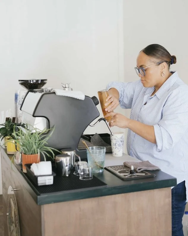 A woman prepares a coffee drink behind a coffee machine in a cafe or kitchen, with various coffee-making tools and plants on the counter.