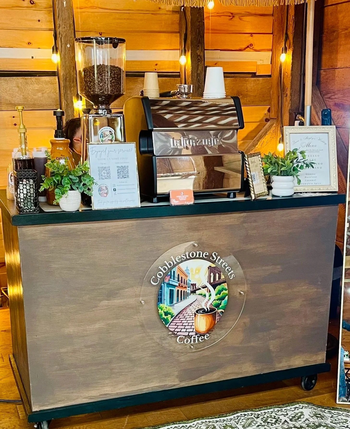 Coffee shop counter with a sign for Cobblestone Streets Coffee, a coffee logo with a colorful street scene, an espresso machine, cups, plant decorations, and a framed menu on a wooden wall background.
