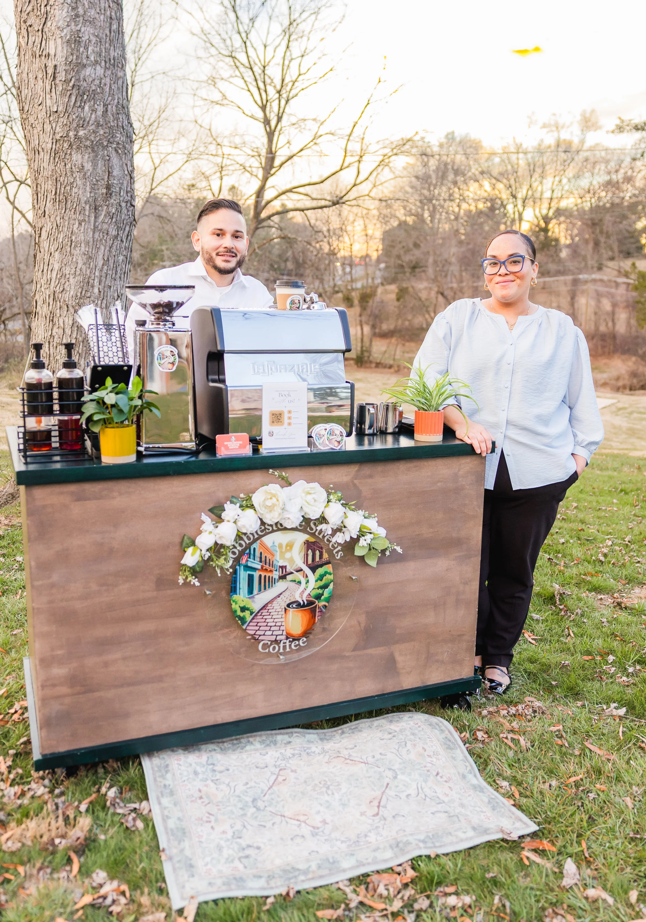 A man and woman standing outdoors behind a wooden coffee stand decorated with flowers, during sunset with trees in the background. The woman has glasses and is smiling, the man is smiling as well, and there is a blanket on the grass in front of them.