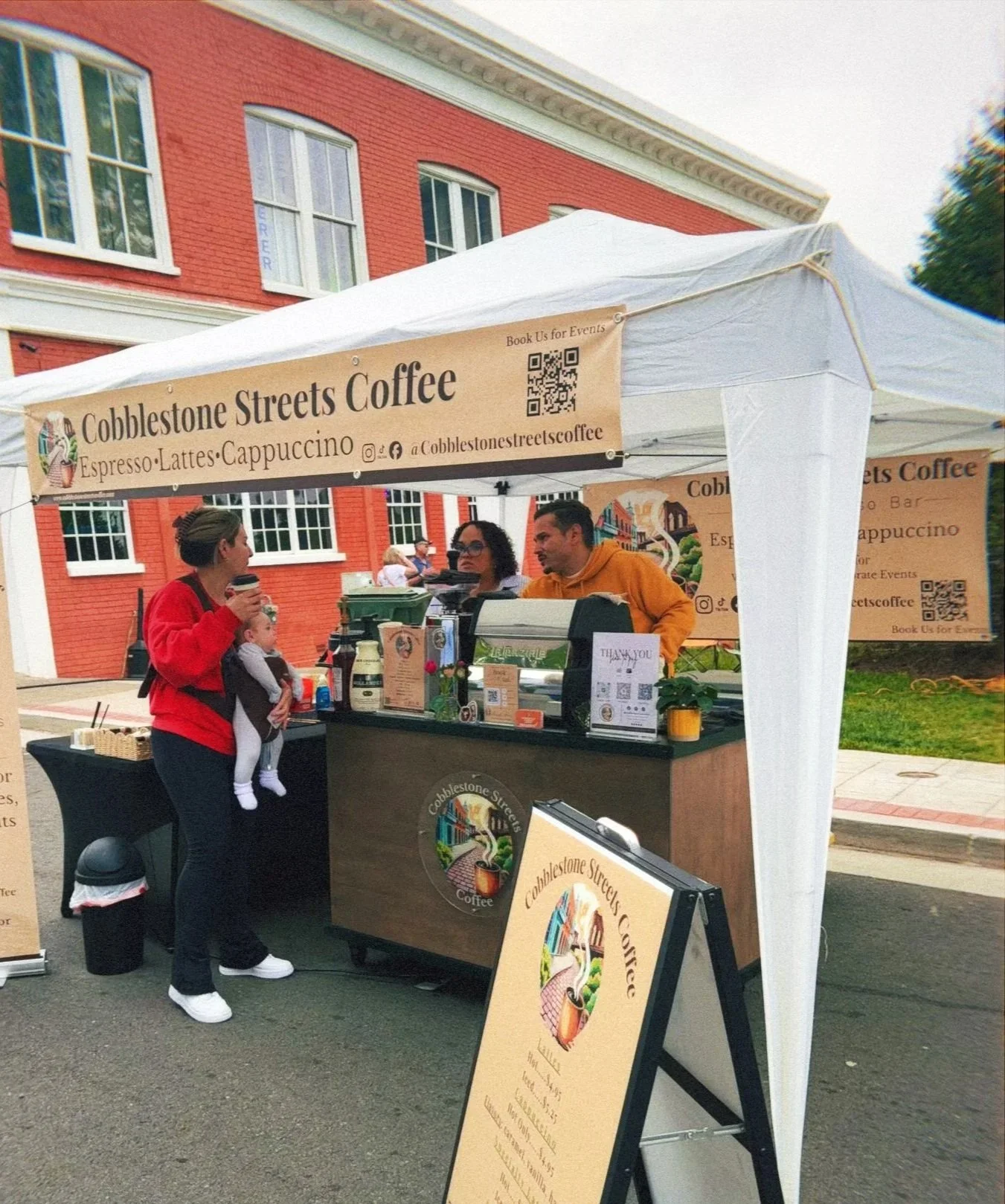 A woman holding a child and a coffee cup at a coffee stand named Cobblestone Streets Coffee, with two staff members behind the counter, in front of a red brick building.