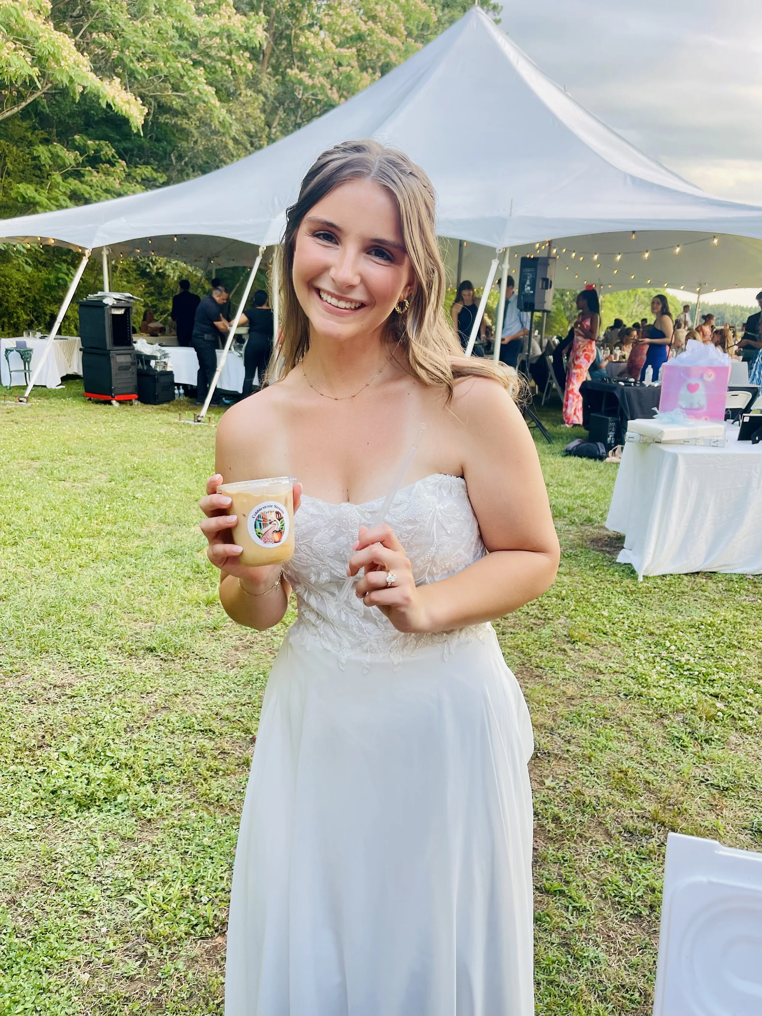 A smiling woman in a white dress holding a dessert cup is at an outdoor event under a large white tent with string lights, surrounded by greenery and people in the background.