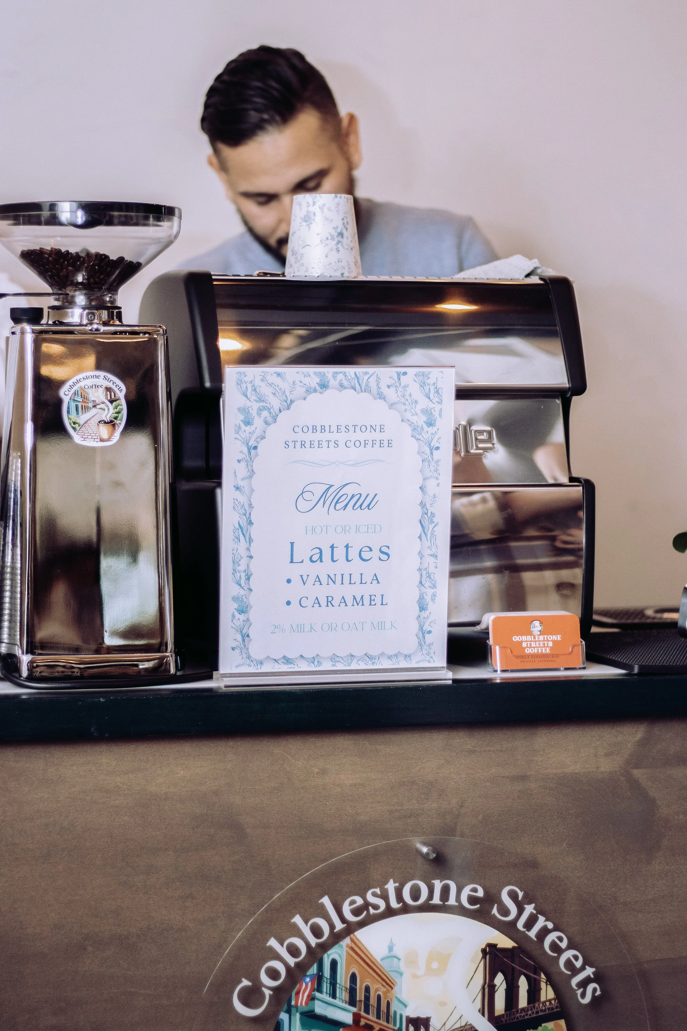 A barista prepares coffee behind a counter with a coffee grinder, a sign with the menu for Cobblestone Streets Coffee, and a metallic coffee machine. The menu lists latte options.