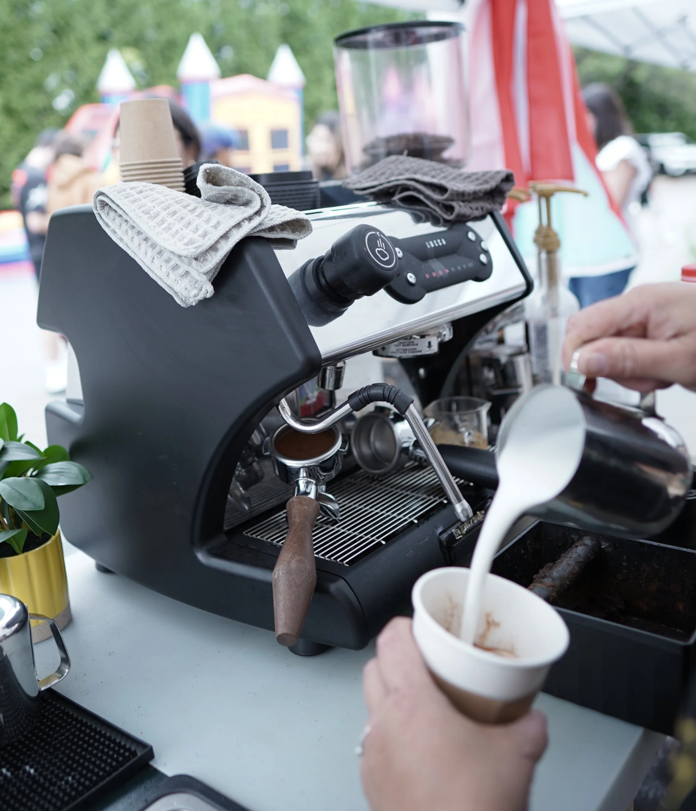Close-up view of a coffee machine, with a barista holding a cup with iced coffee, in an outdoor setting with blurred people in the background.