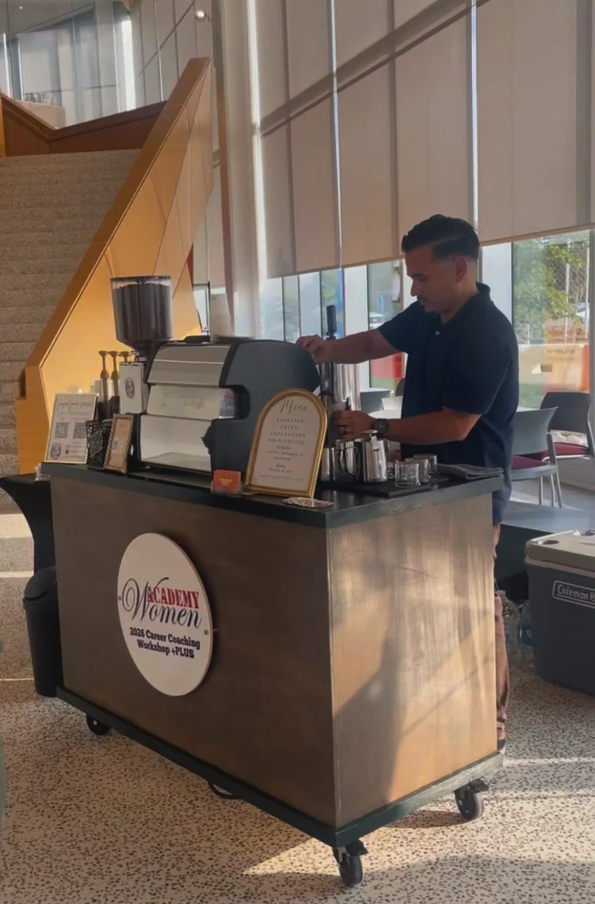 A barista preparing coffee at a mobile coffee cart with a sign that reads 'Academy Women 2023 Career Coaching Workshop +PLUS' in a spacious indoor setting with large windows and natural light.