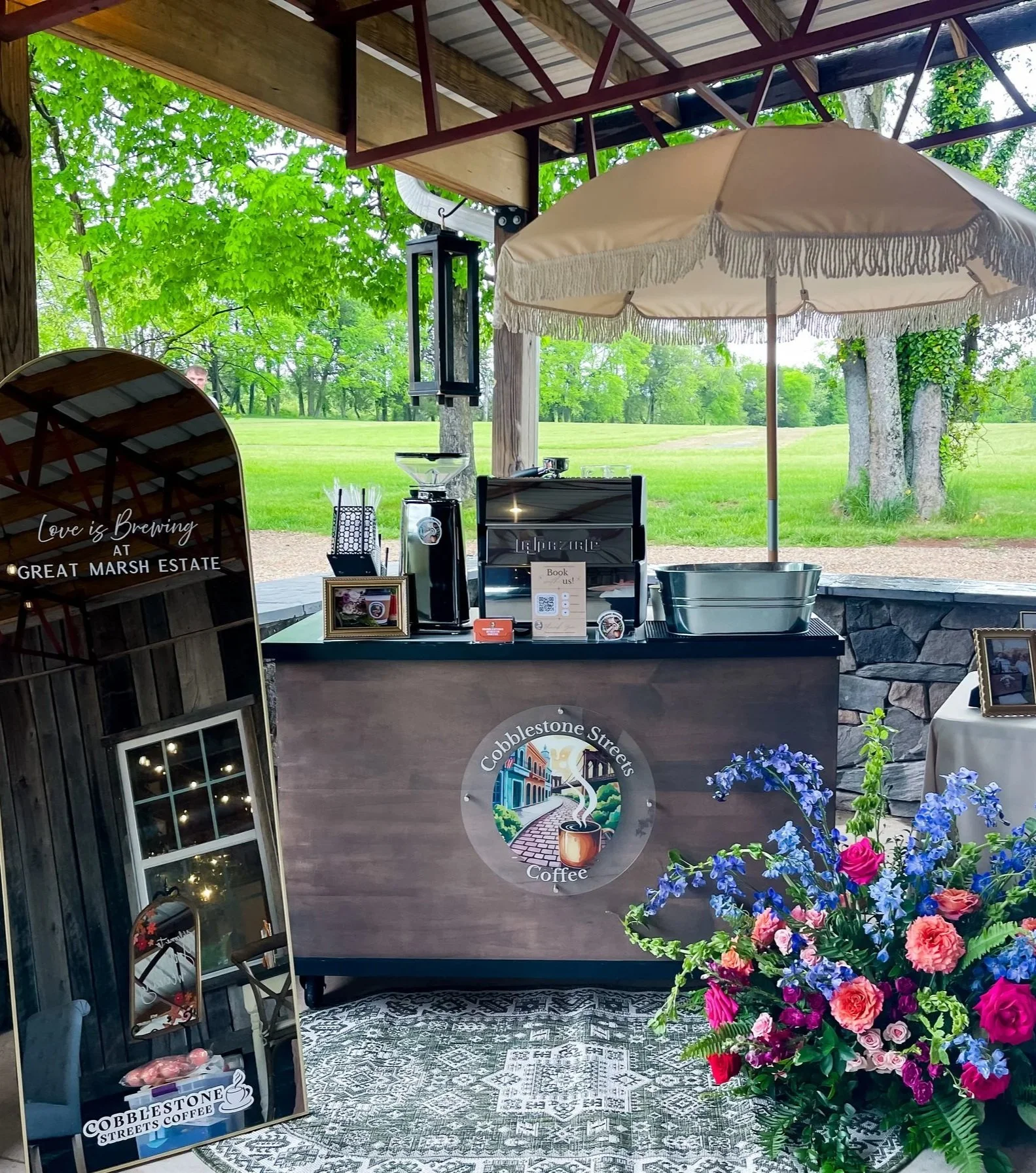 Outdoor coffee stand with a large beige patio umbrella, a black counter with coffee equipment, a floral arrangement, and a sign that reads 'Love is brewing at Cobblestone Streets Coffee', set in a lush green park.