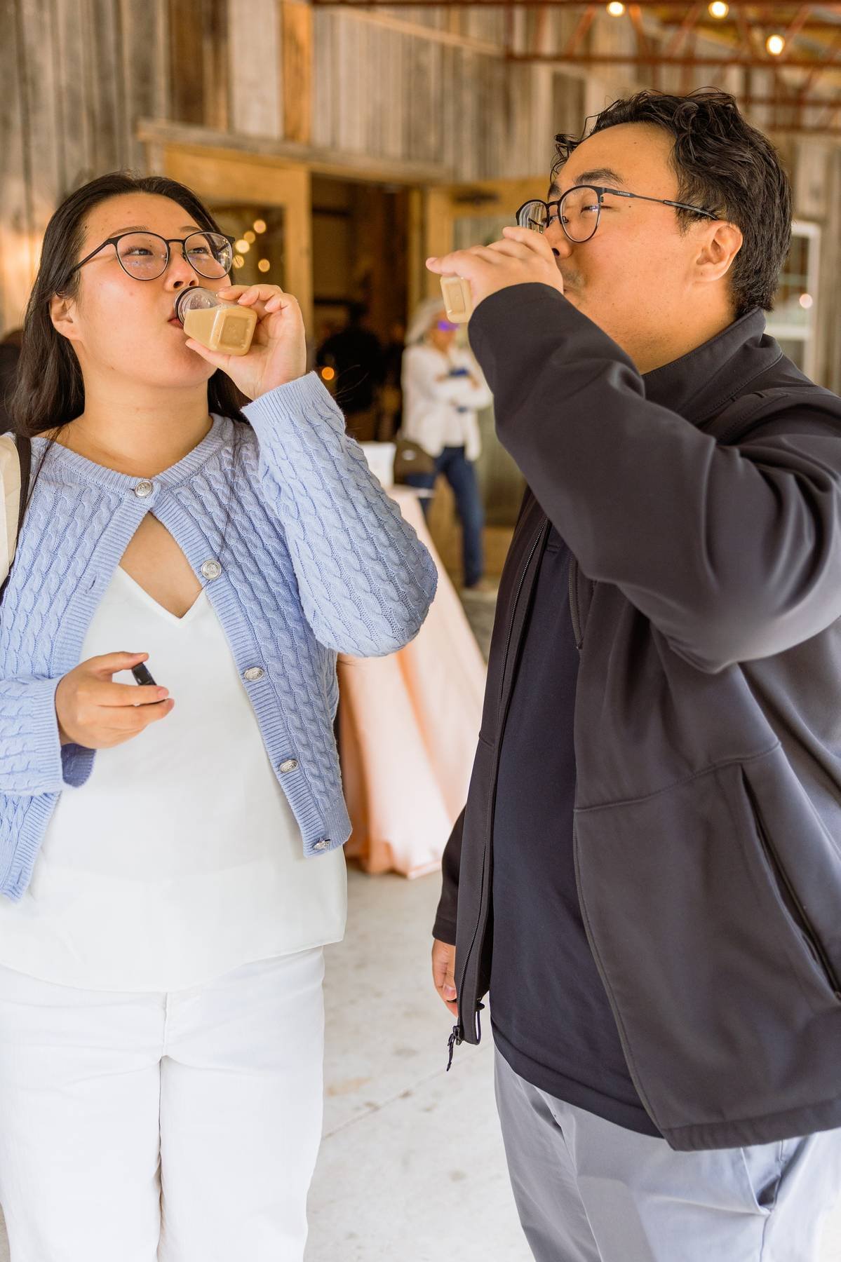 Two people drinking coffee in a rustic indoor setting with string lights, one woman wearing glasses and a light blue cardigan, and a man wearing glasses and a black jacket.