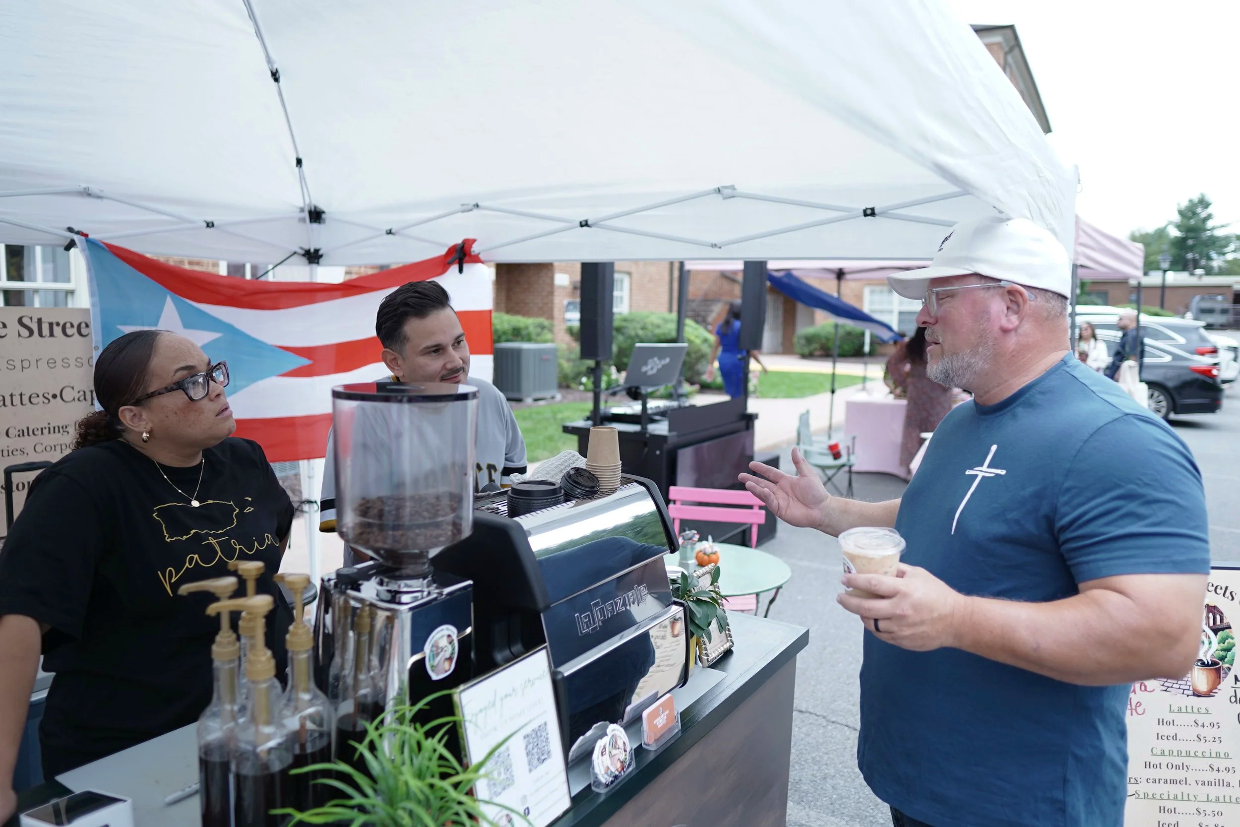 People at an outdoor café or market stand, engaging in conversation with coffee and menu signs displayed on the stand, with a Puerto Rican flag in the background.