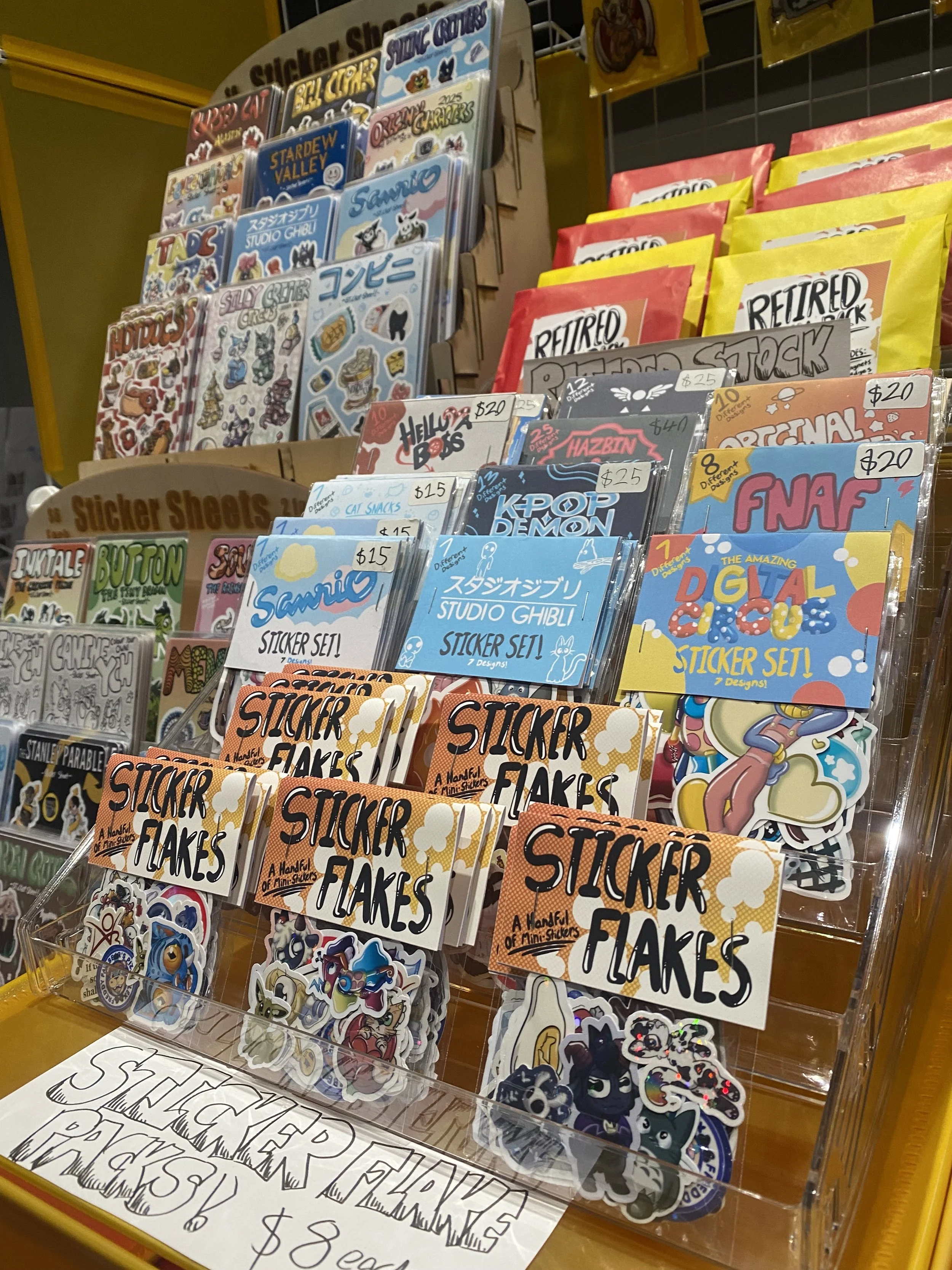 Display of various sticker sheets featuring characters from Studio Ghibli movies and other designs, arranged on a stand. Price tags and labels indicate they are mini sticker sets, with some priced at $20 and $25.