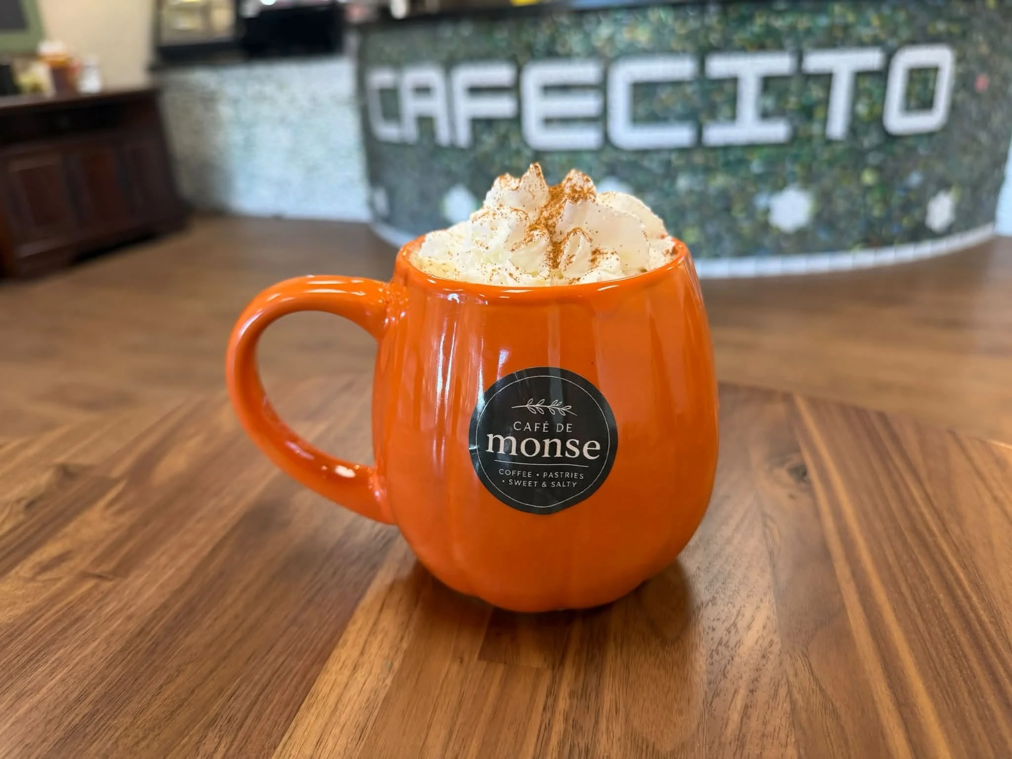 A bright orange mug filled with whipped cream and sprinkled with cinnamon, placed on a wooden table in a coffee shop with a blurred background showing a wall with the word 'CHELLO'.