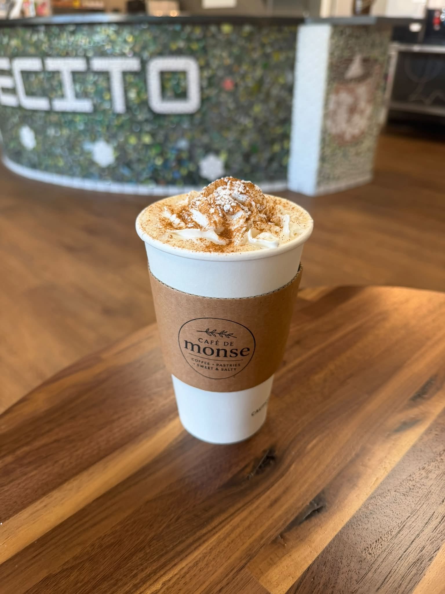 A takeout coffee cup at Café de Monse with whipped cream, dusted with cinnamon or cocoa powder, on a wooden table in a coffee shop.