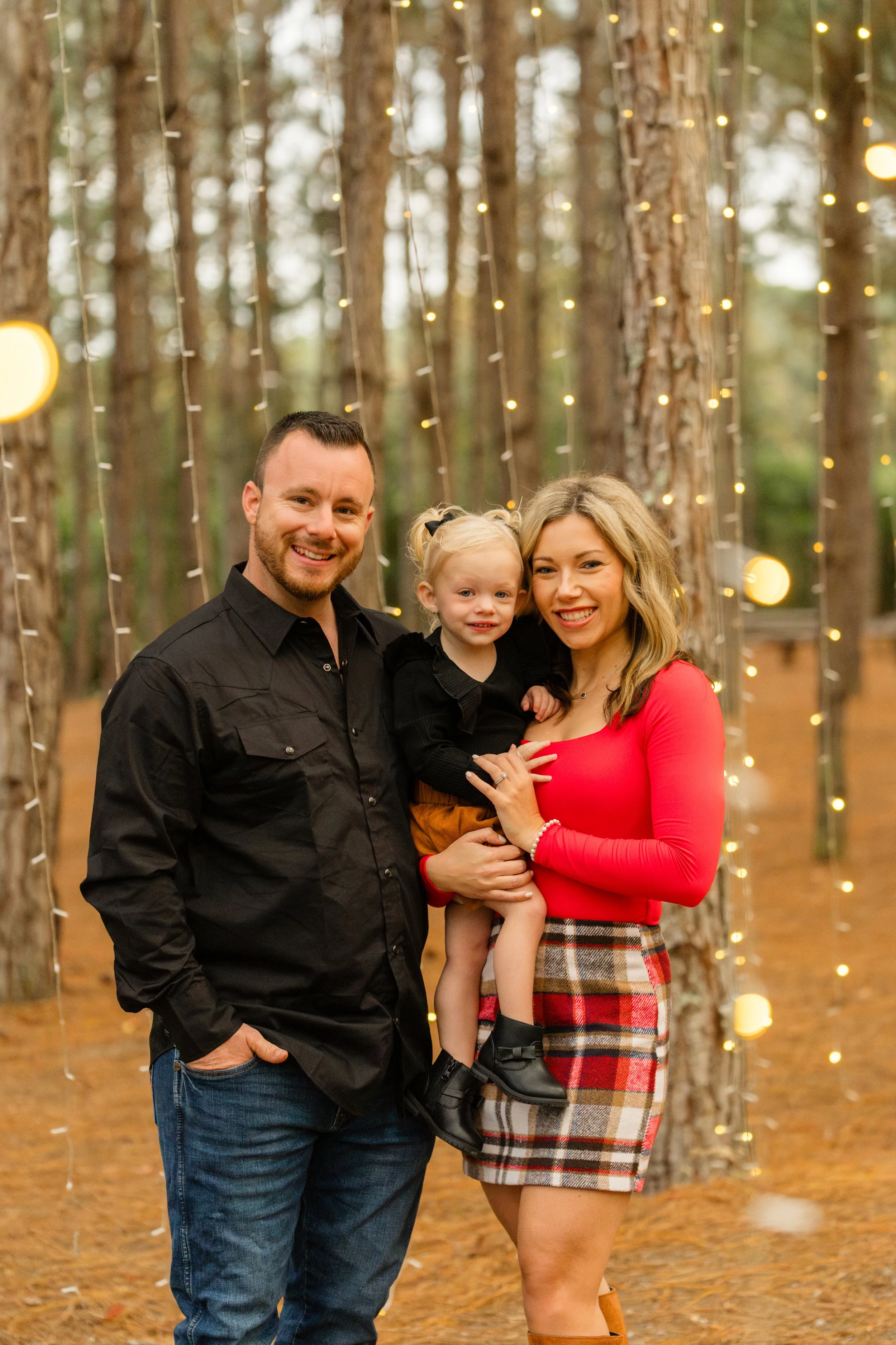 A family of three smiling for a photo in a wooded area decorated with string lights during the evening. The man is wearing a black shirt, the woman is in a red top with plaid skirt, and the young girl is in a black top with black boots.