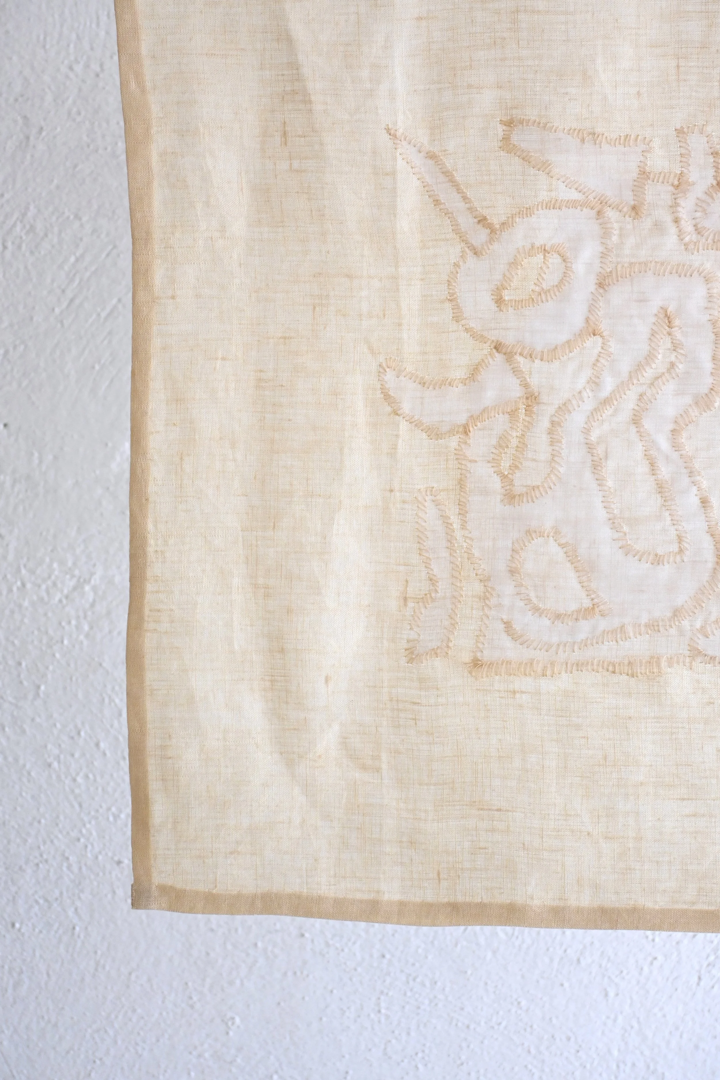 Close-up of a beige fabric curtain with embroidered floral design, hanging on a white textured wall.