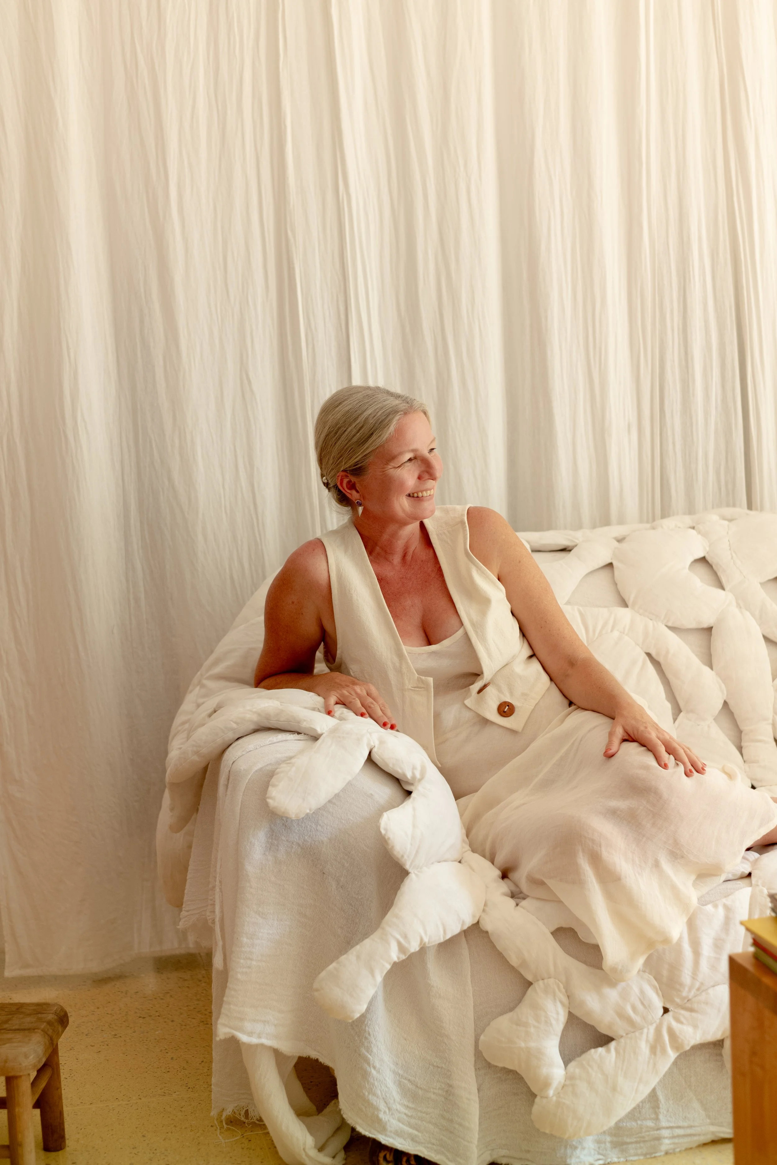 A smiling woman with gray hair sitting on a bed surrounded by stuffed animals, in a room with cream-colored curtains.