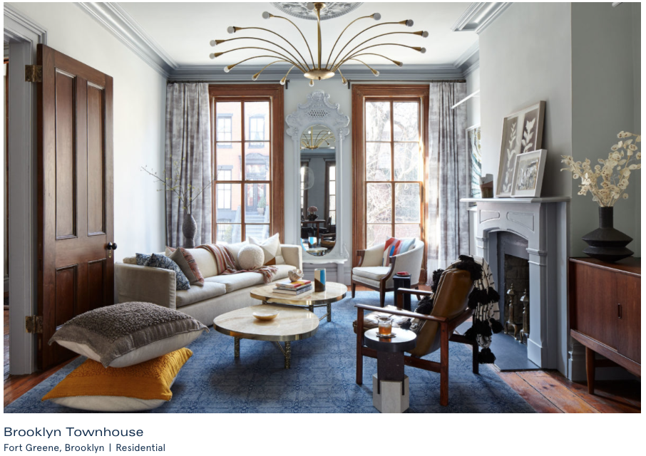 A cozy living room with a beige sofa, two round coffee tables, and various armchairs. The room features large windows with curtains, a decorative mirror, a fireplace, and a wooden cabinet. There are pillows and a blanket on the furniture, and decorat