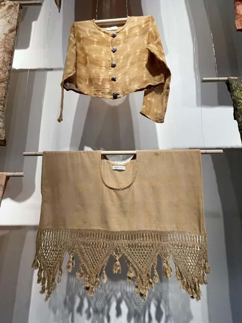 Two beige fabrics hanging on rods in a store display. The top fabric is a cropped jacket with button-down front and rough texture. The bottom fabric is a decorative shawl or scarf with intricate crochet lace and tassel fringe.