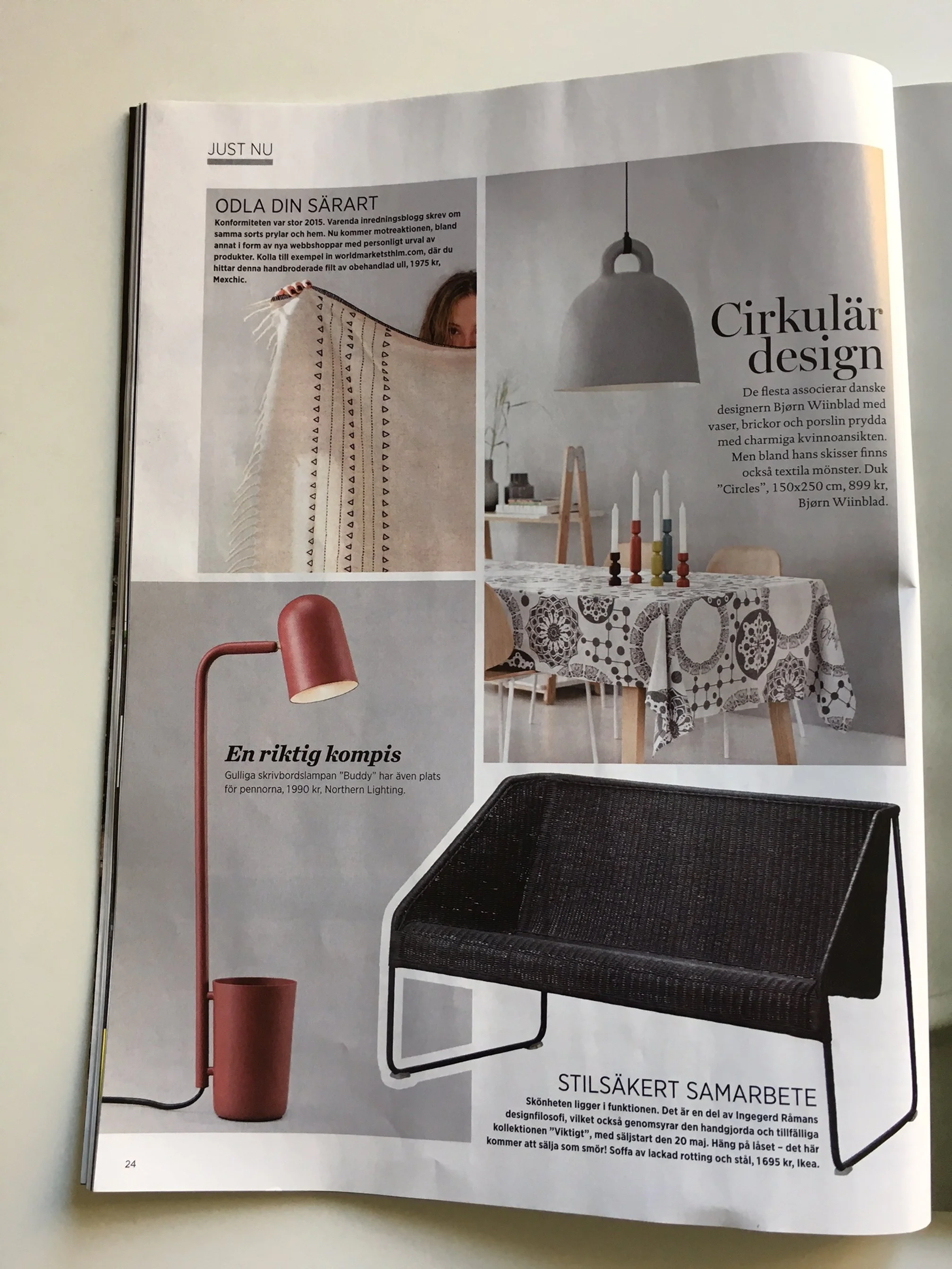 Open magazine page showing a black rattan sofa, a pink floor lamp, and a round table with a floral tablecloth, candles, and decorative objects in a modern interior.