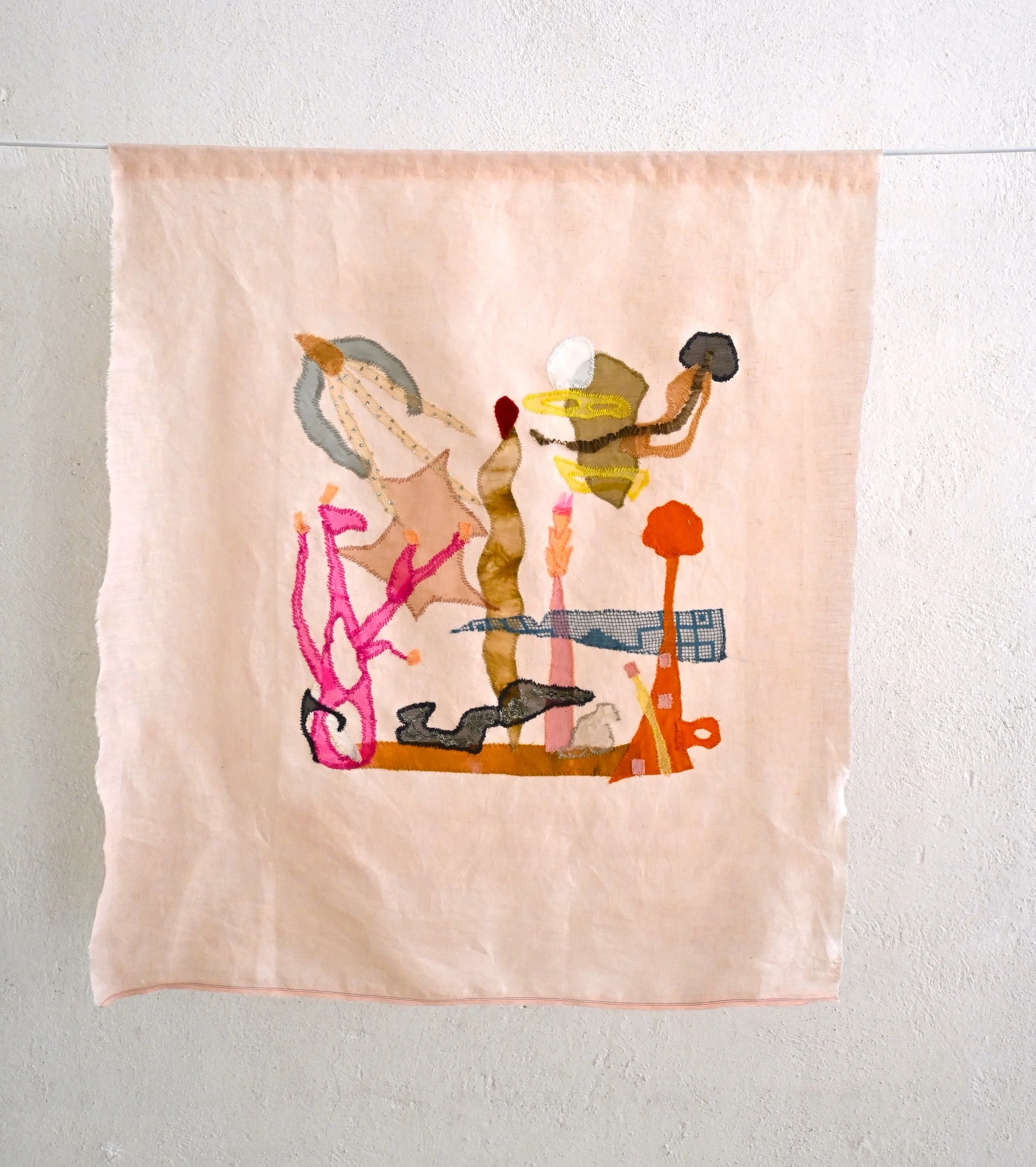 A piece of pink fabric with a colorful embroidered design hanging on a horizontal rod against a white textured wall.