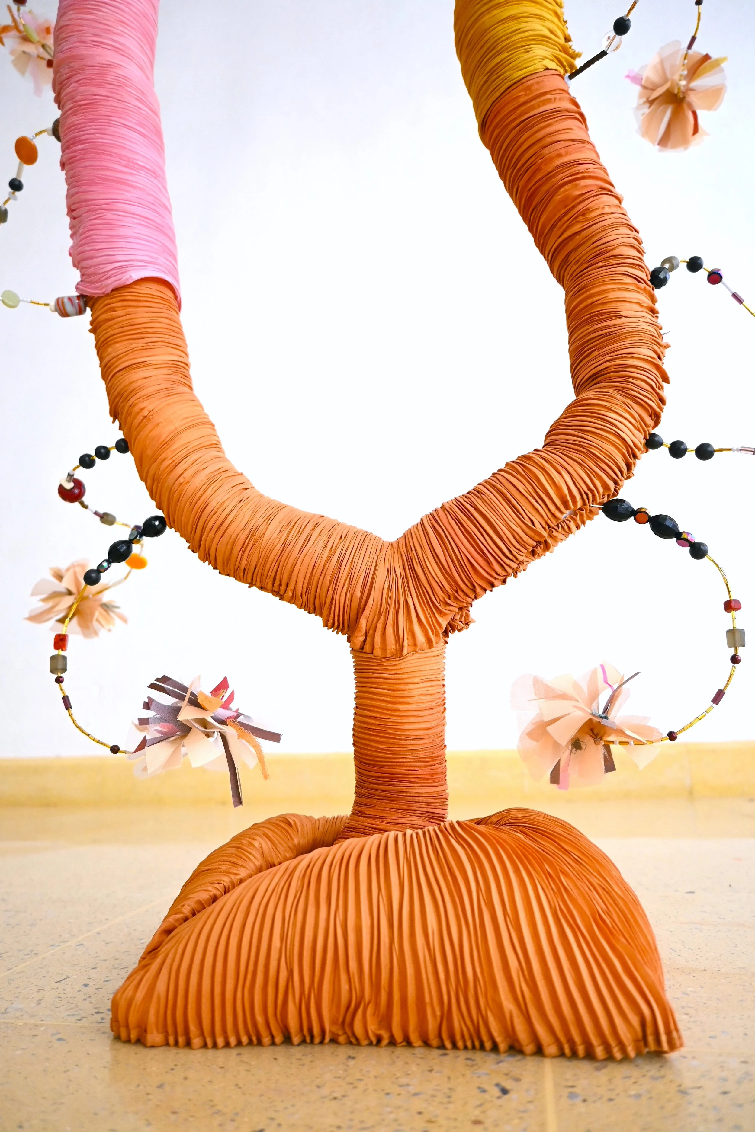 A colorful handmade sculpture resembling a tree with a large orange base, a twisted brown trunk, and branches wrapped in pink and orange fabric, decorated with paper flowers and beaded garlands.
