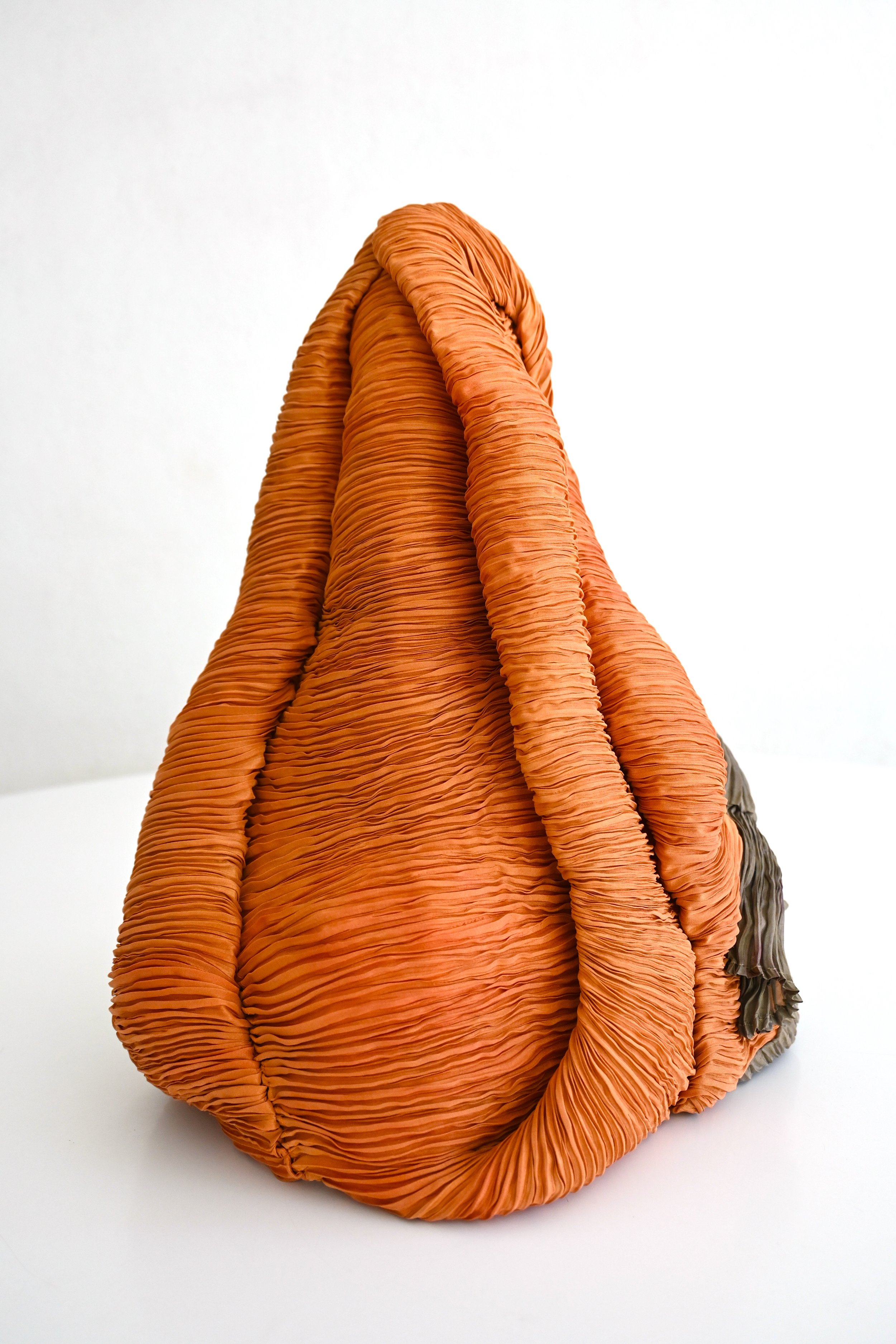 A large, layered, orange fabric sculpture resembling a coiled, wrinkled, organic form, set against a white background.