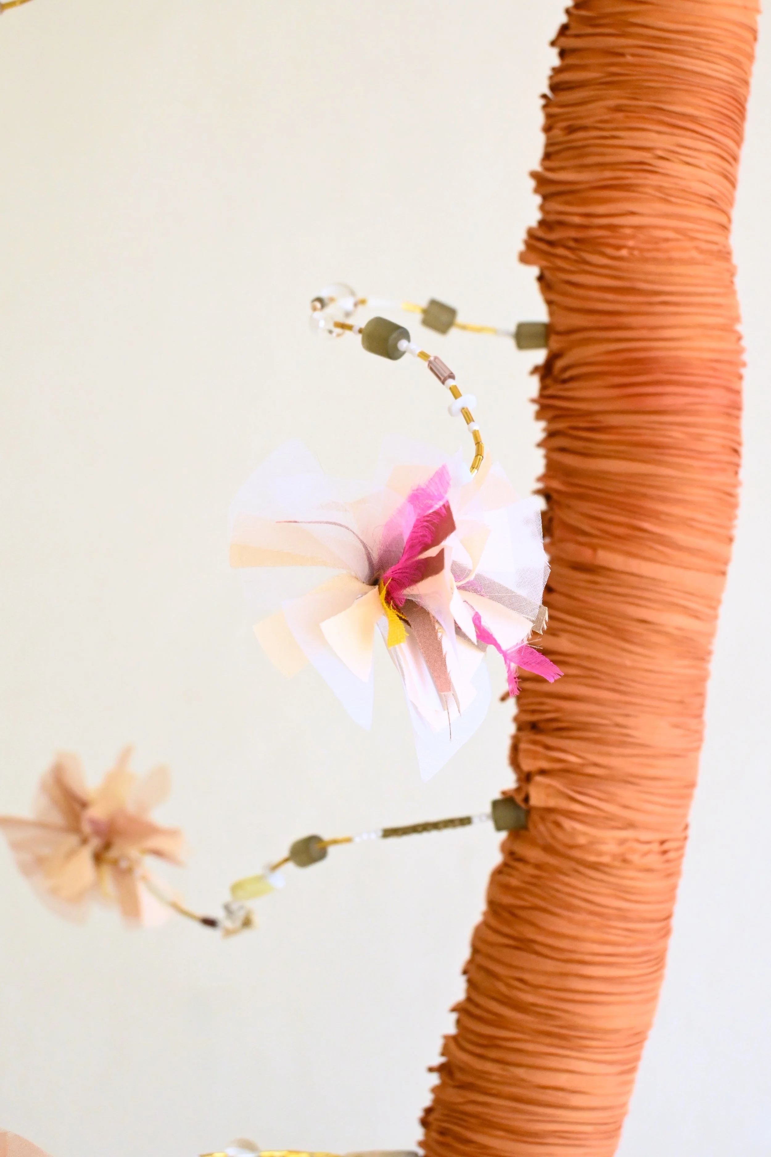 A close-up of a decorative arrangement featuring a tall, orange, rolled-paper column and a branch with a paper flower and beads attached to it.