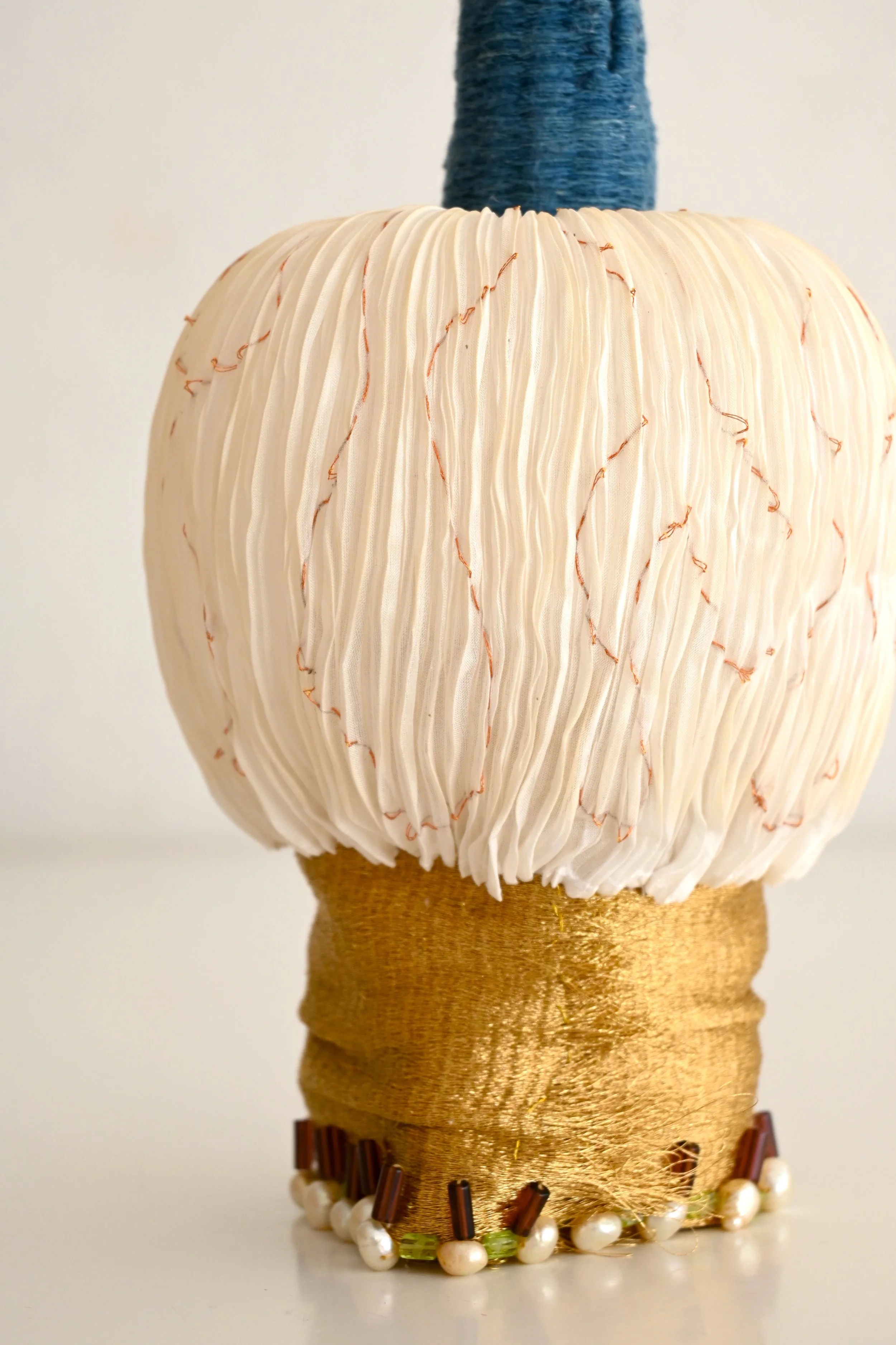 Fashion mannequin head with a short, pleated, light pink and cream-colored wig, decorated with orange thread accents, set on a golden base, surrounded by artificial pearls, and topped with a blue, textured hat or head covering.