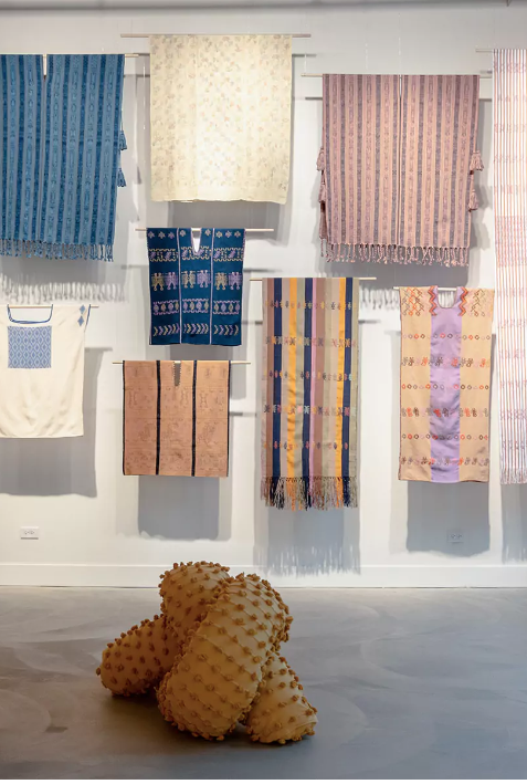 Colorful woven and fabric art pieces hanging on a wall with a textured, plush sculpture on the floor.