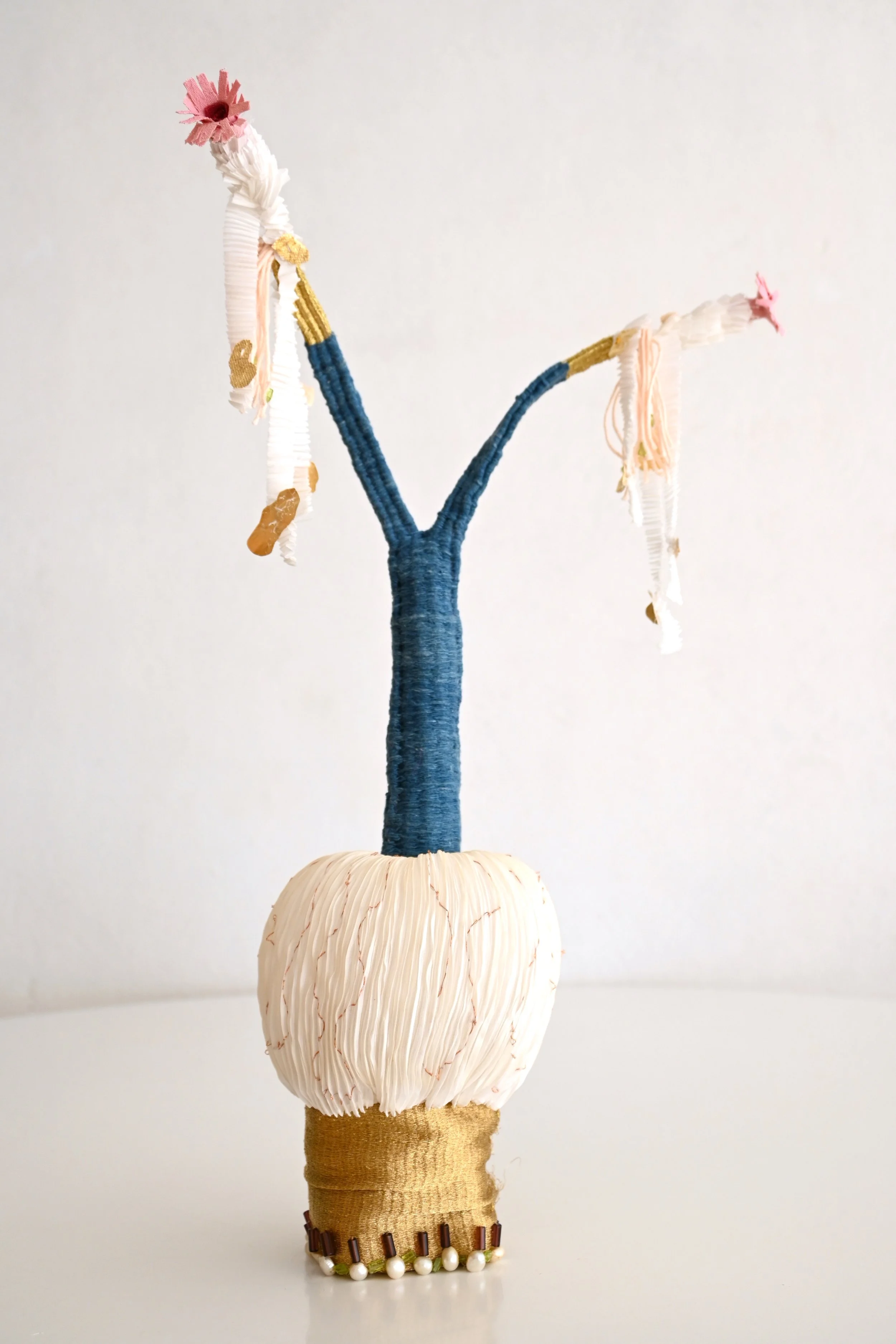 An art piece resembling a bare tree made from various craft materials, with a white, rounded base, a textured white branch, and two branches wrapped in blue and white cloth, topped with pink flowers, set against a plain white background.