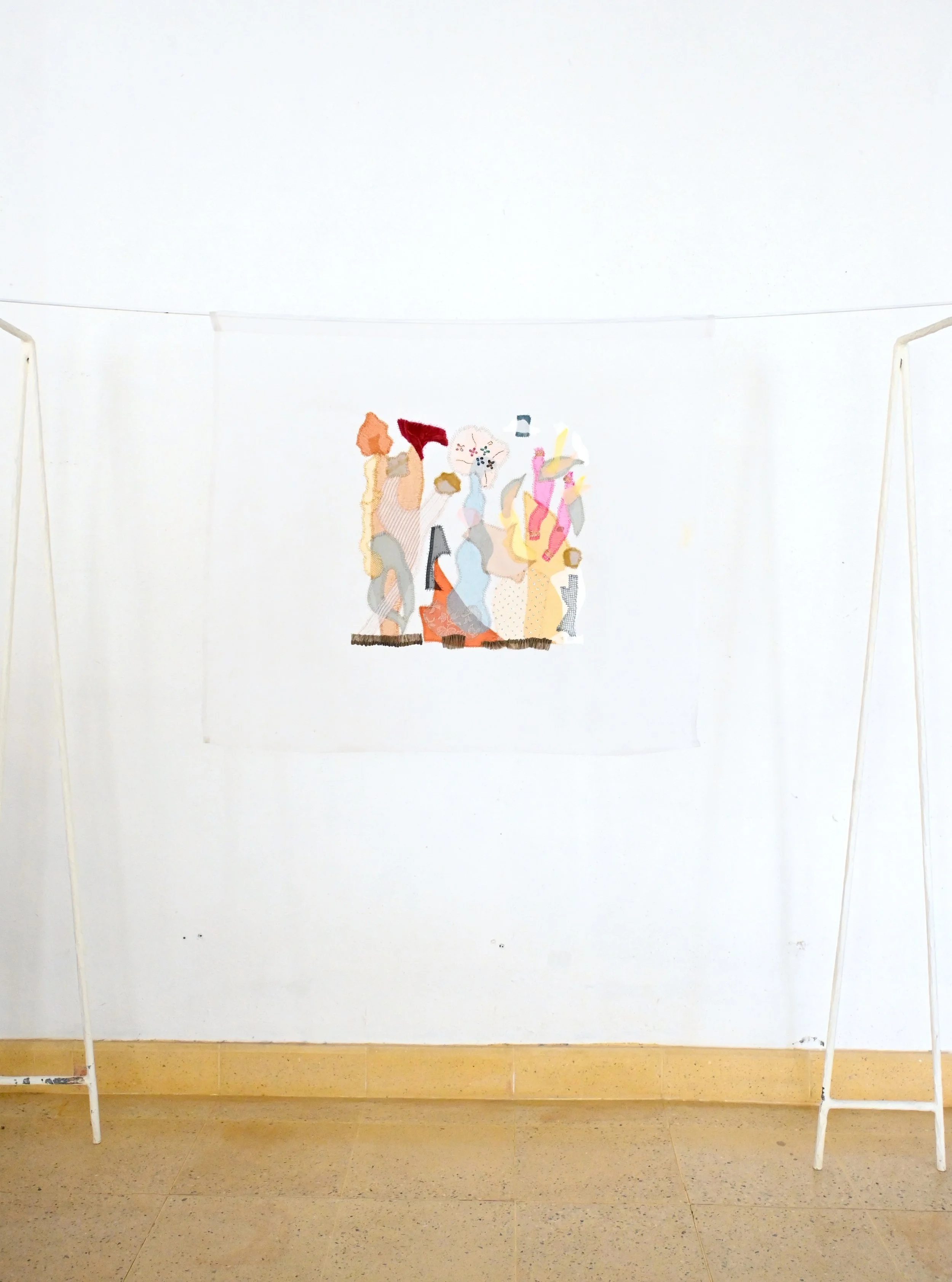 A white backdrop with a colorful embroidered textile artwork hanging from a thin wire. The artwork depicts abstract shapes and figures in pastel and bright colors, mounted on a plain white wall. There are two white freestanding supports on either side of the backdrop.