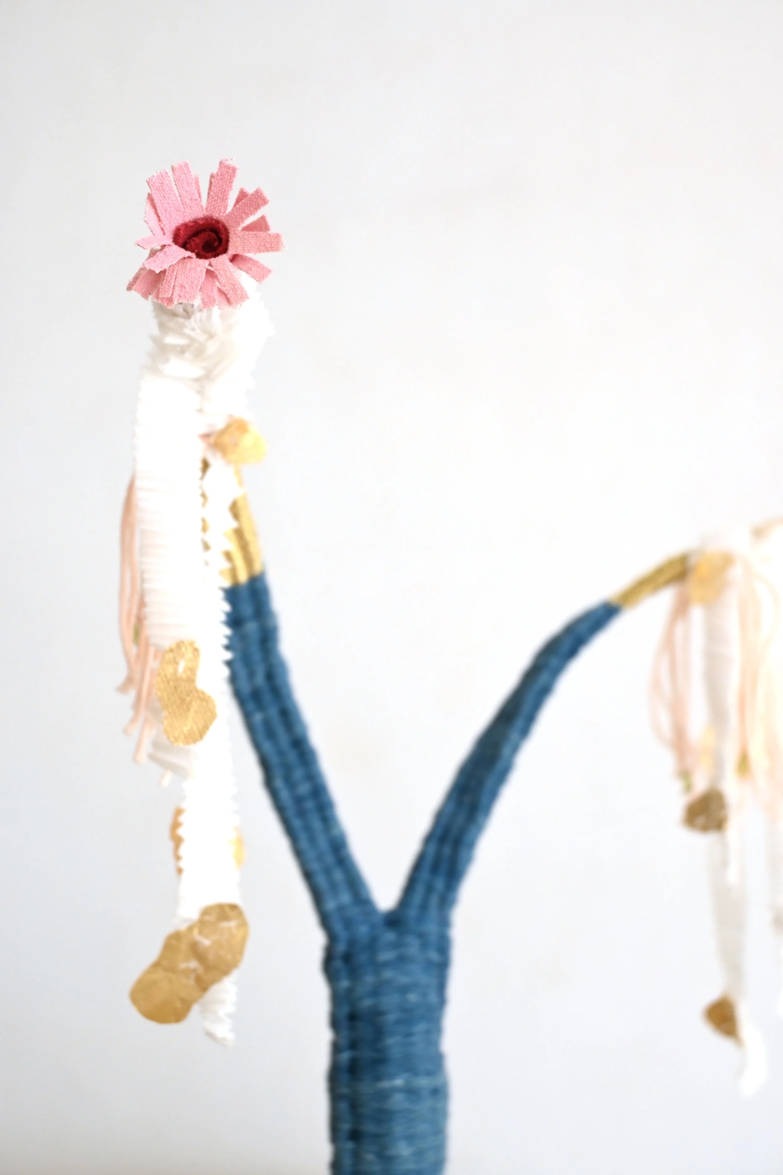 Close-up of a handmade craft object resembling a minimalist flower with pink fabric petals and a dark red center, on a blue textured stem against a plain light background.