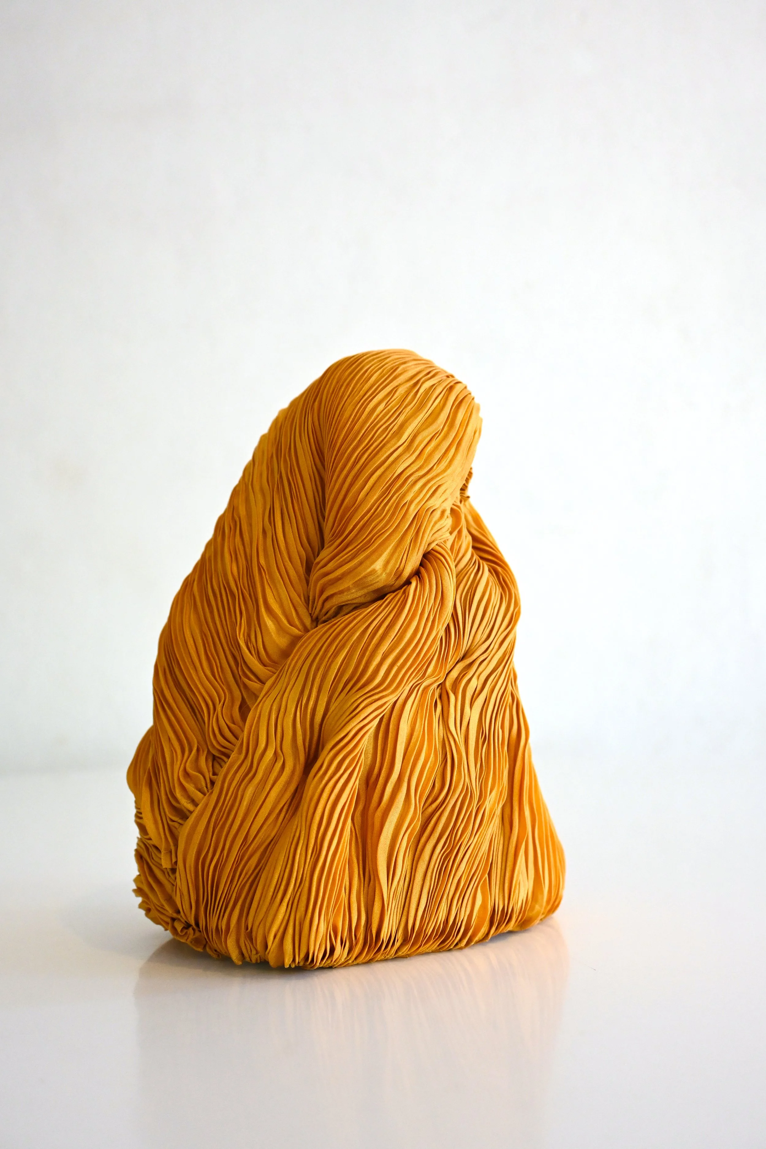 A large, orange, textured cloth sculpture resembling a draped garment or abstract form sits on a white surface against a plain white background.