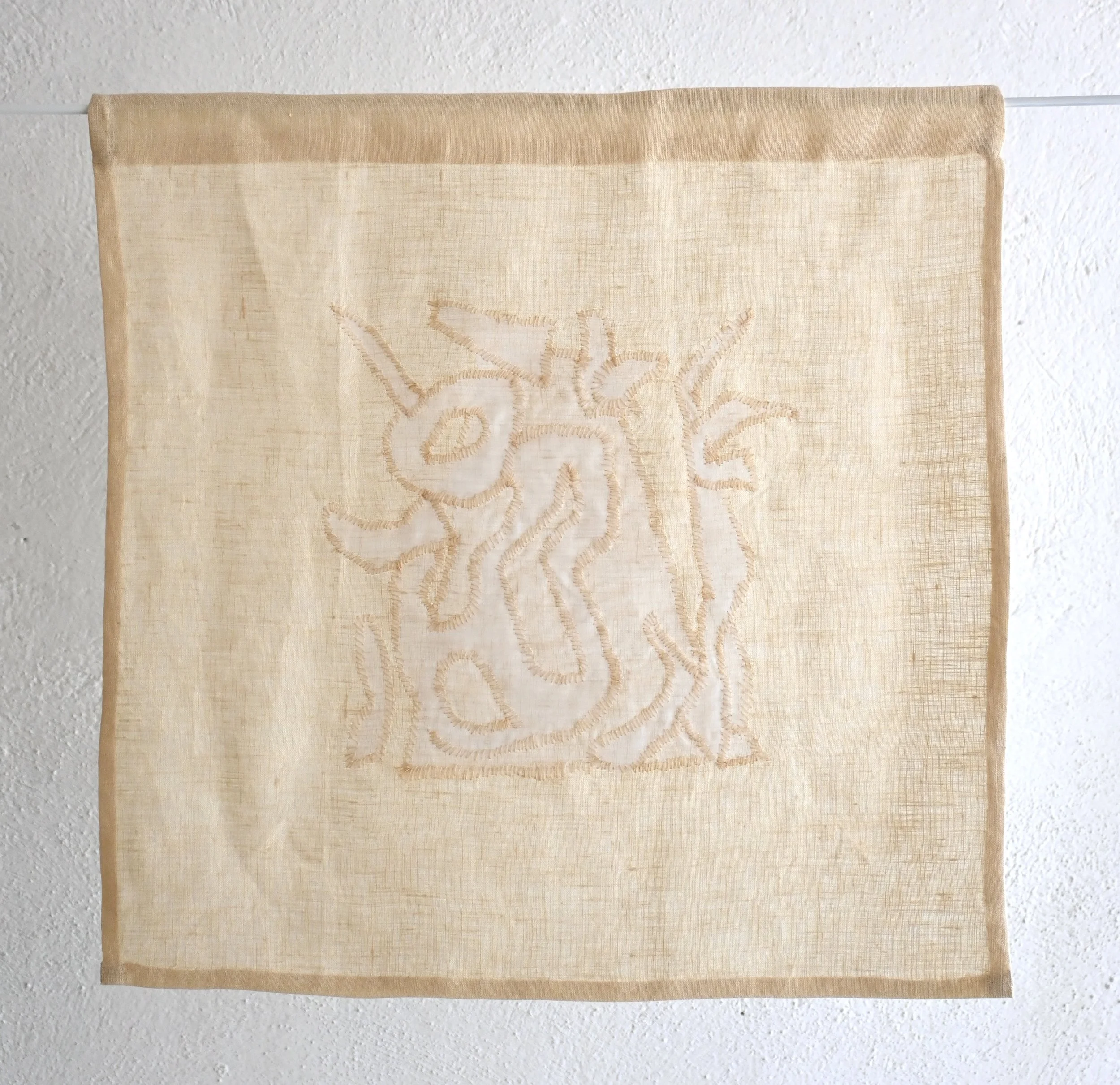Close-up of a beige embroidered fabric banner hanging on a white wall.