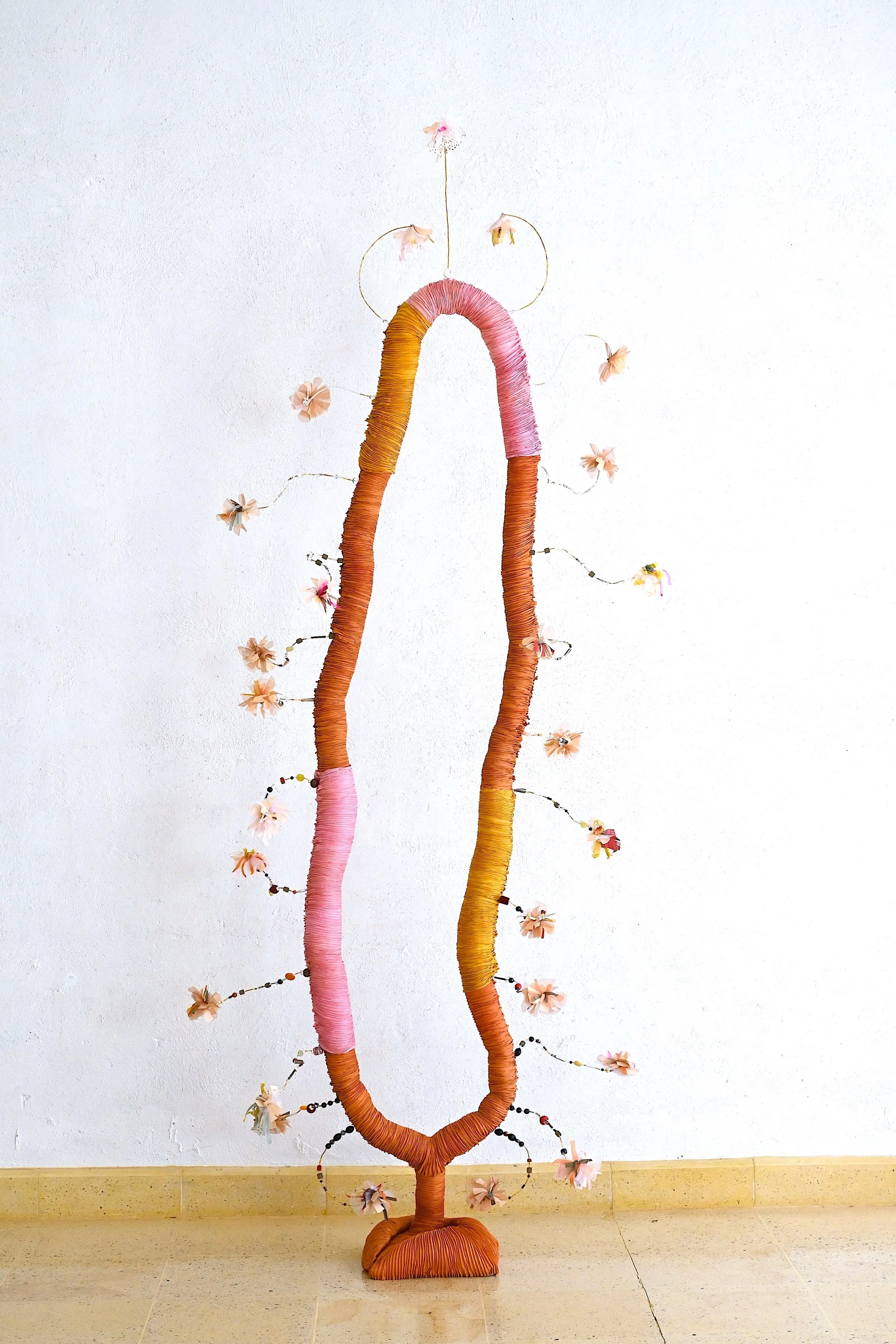 A decorative art piece resembling two elongated, curved shapes wrapped in orange and pink fabric, standing vertically against a white wall. The shapes are adorned with small, artificial flowers and connected by thin wire.
