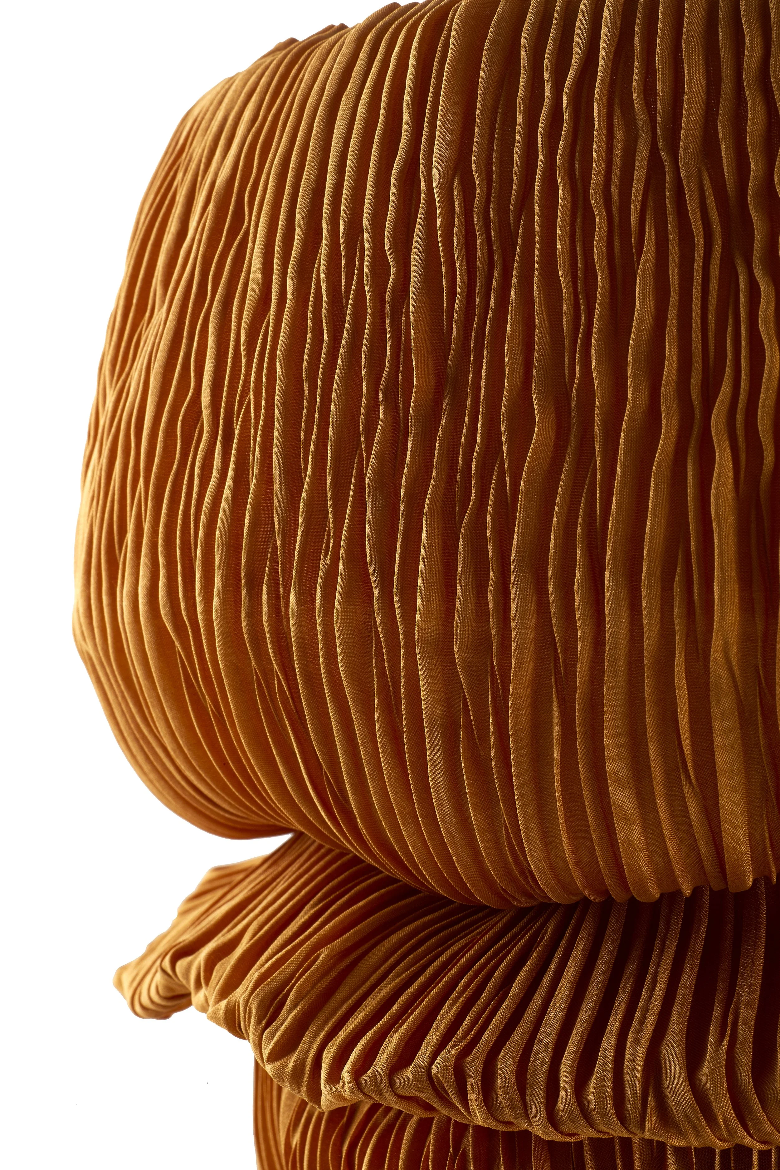 Close-up of a pleated, orange-brown fabric with a sculptural, rounded shape, reflected on a glossy surface.
