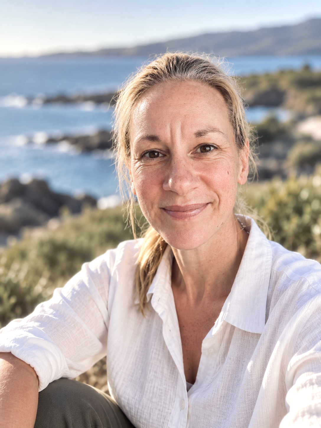 A woman with blonde hair and a white shirt smiling outdoors with a body of water and land in the background.