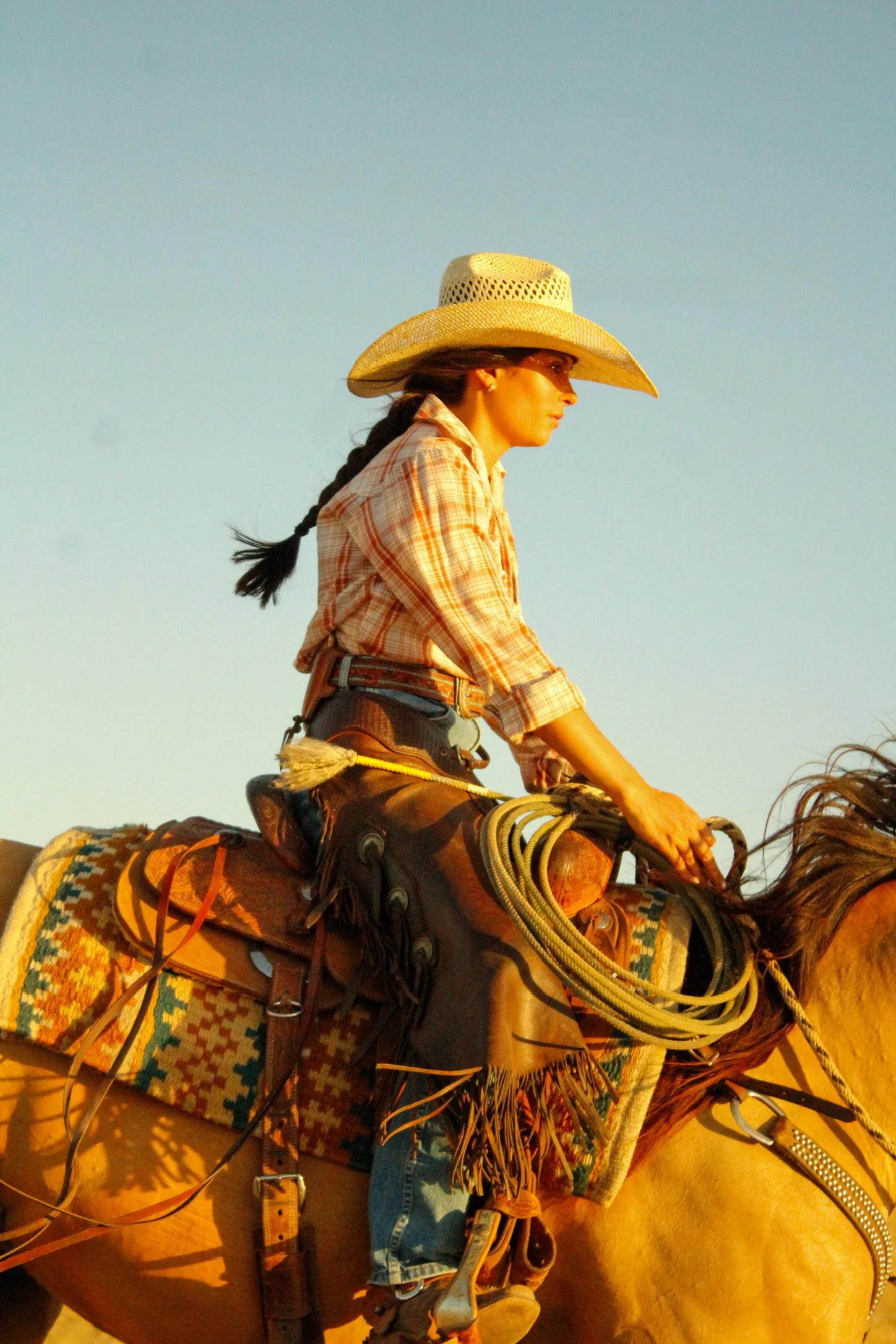 Cowgirl wearing a straw hat and plaid shirt riding a saddled horse outdoors