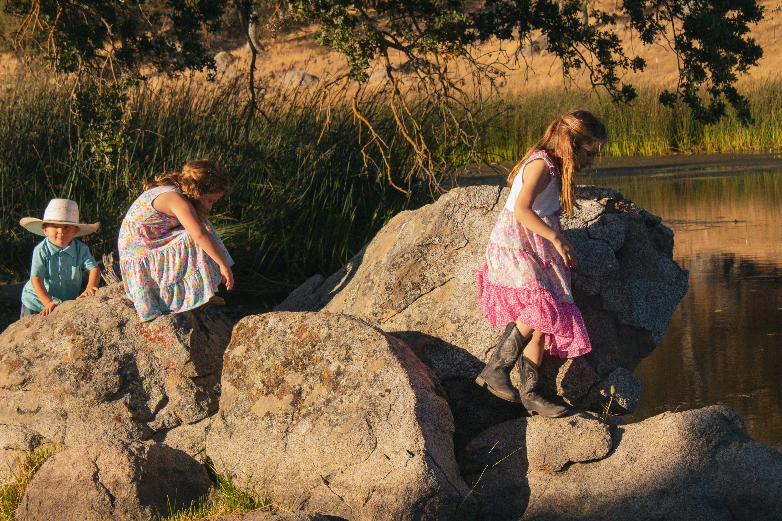 Three children exploring rocks beside a pond, including a boy in a cowboy hat and two girls in dresses.