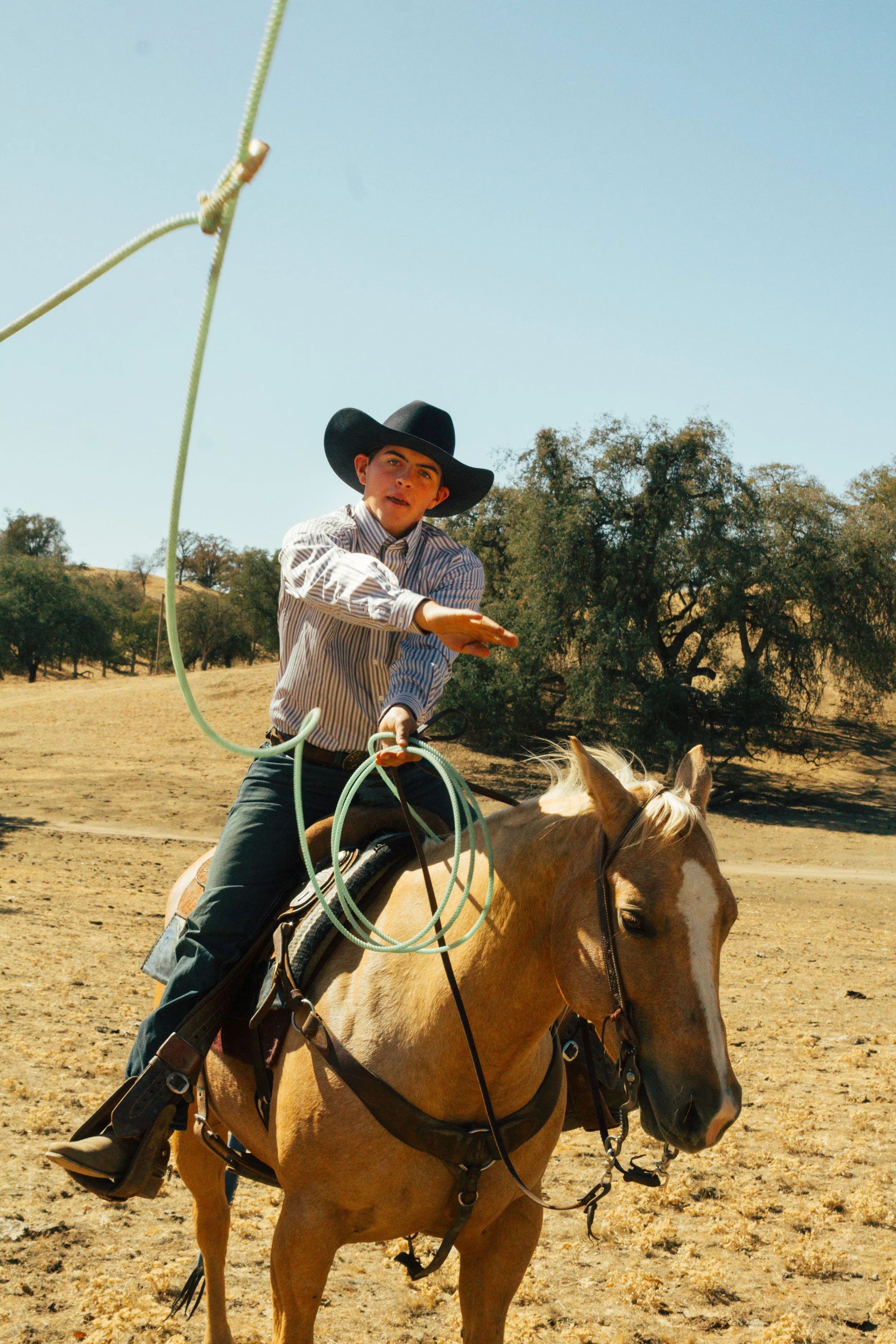 Young cowboy on horseback swinging a lasso in a dry rural field
