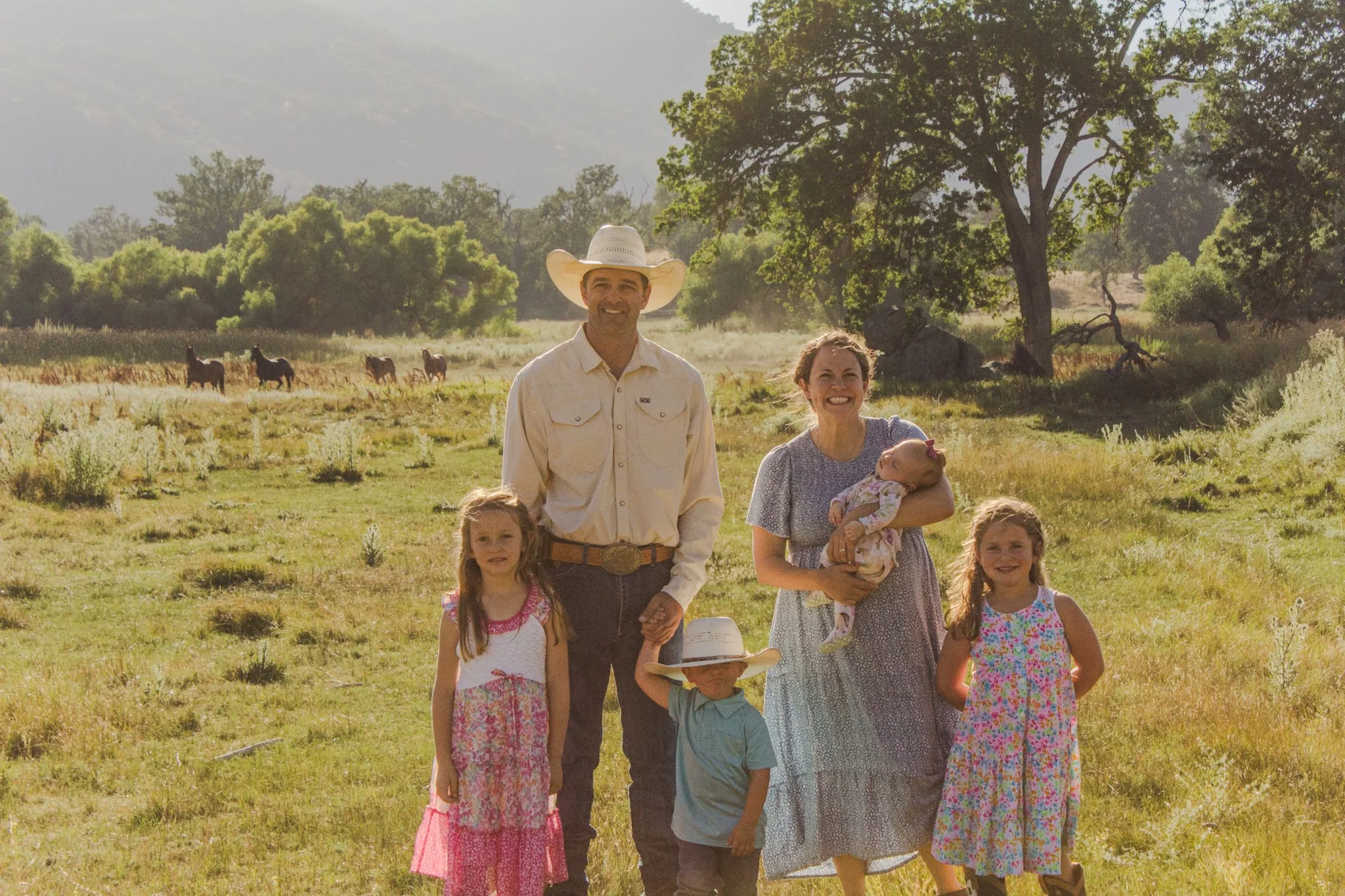 Family posing in a sunny meadow with horses, trees, and mountains in the background
