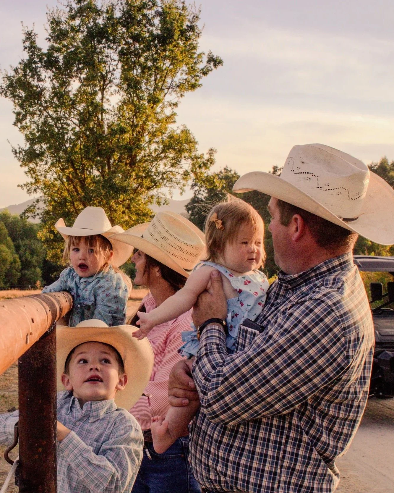 Family wearing cowboy hats gathers outdoors at sunset with three young children near a ranch fence.