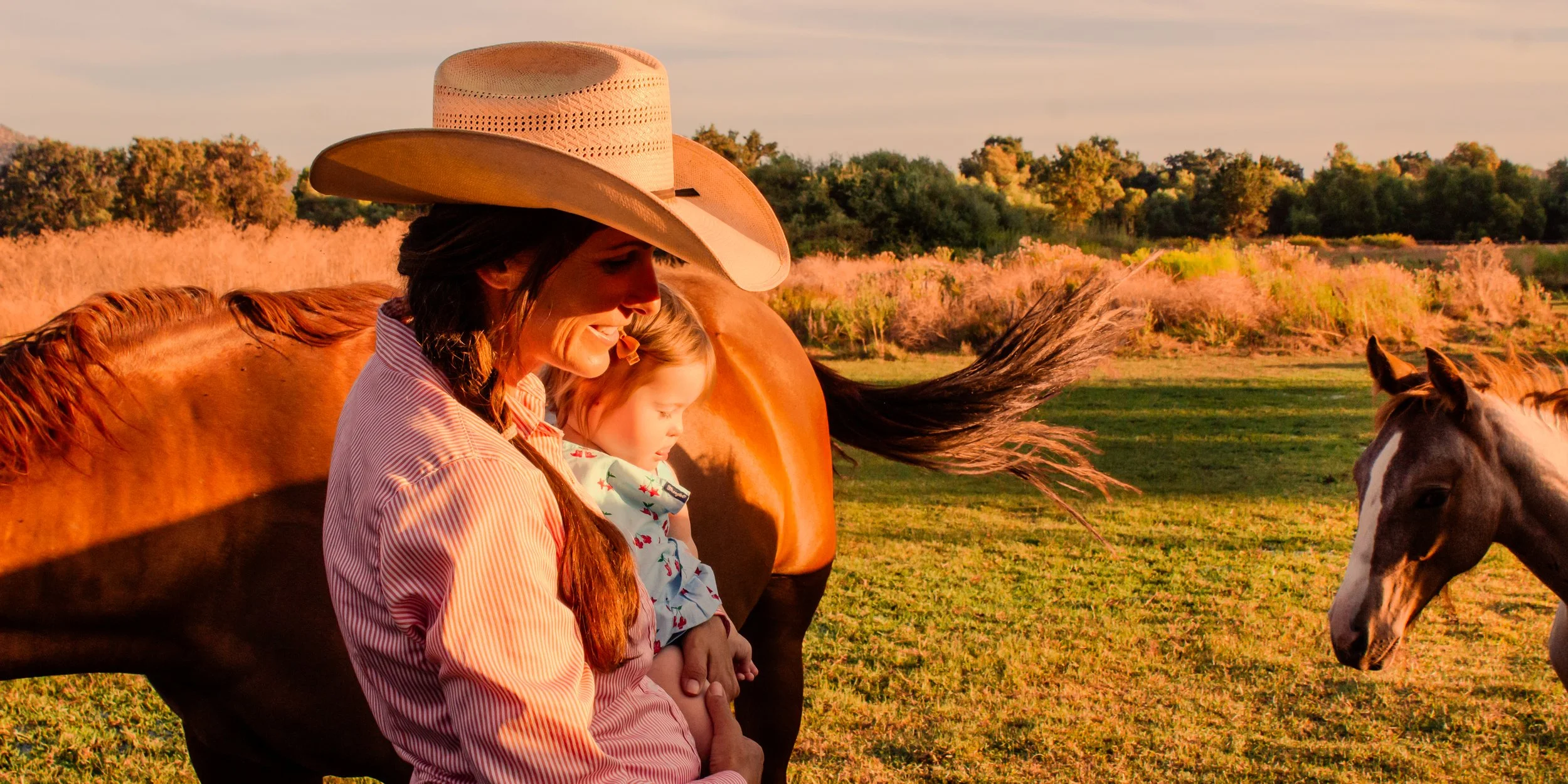 Woman in a cowboy hat holding a child beside horses in a sunlit field