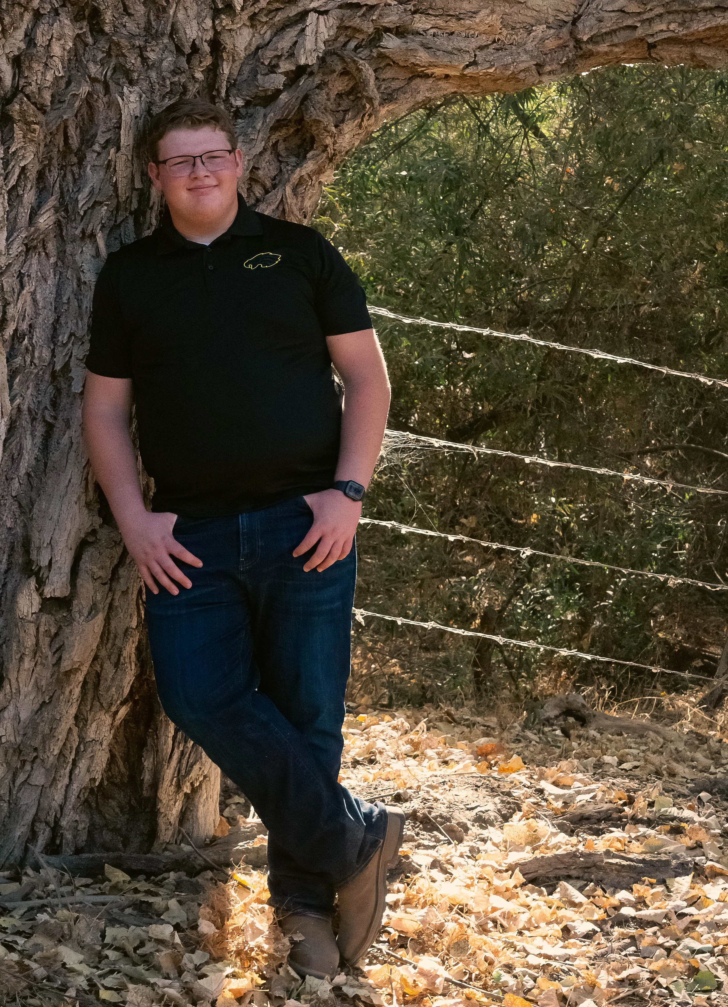Young man wearing glasses and a black polo shirt standing beside a large tree in a wooded area
