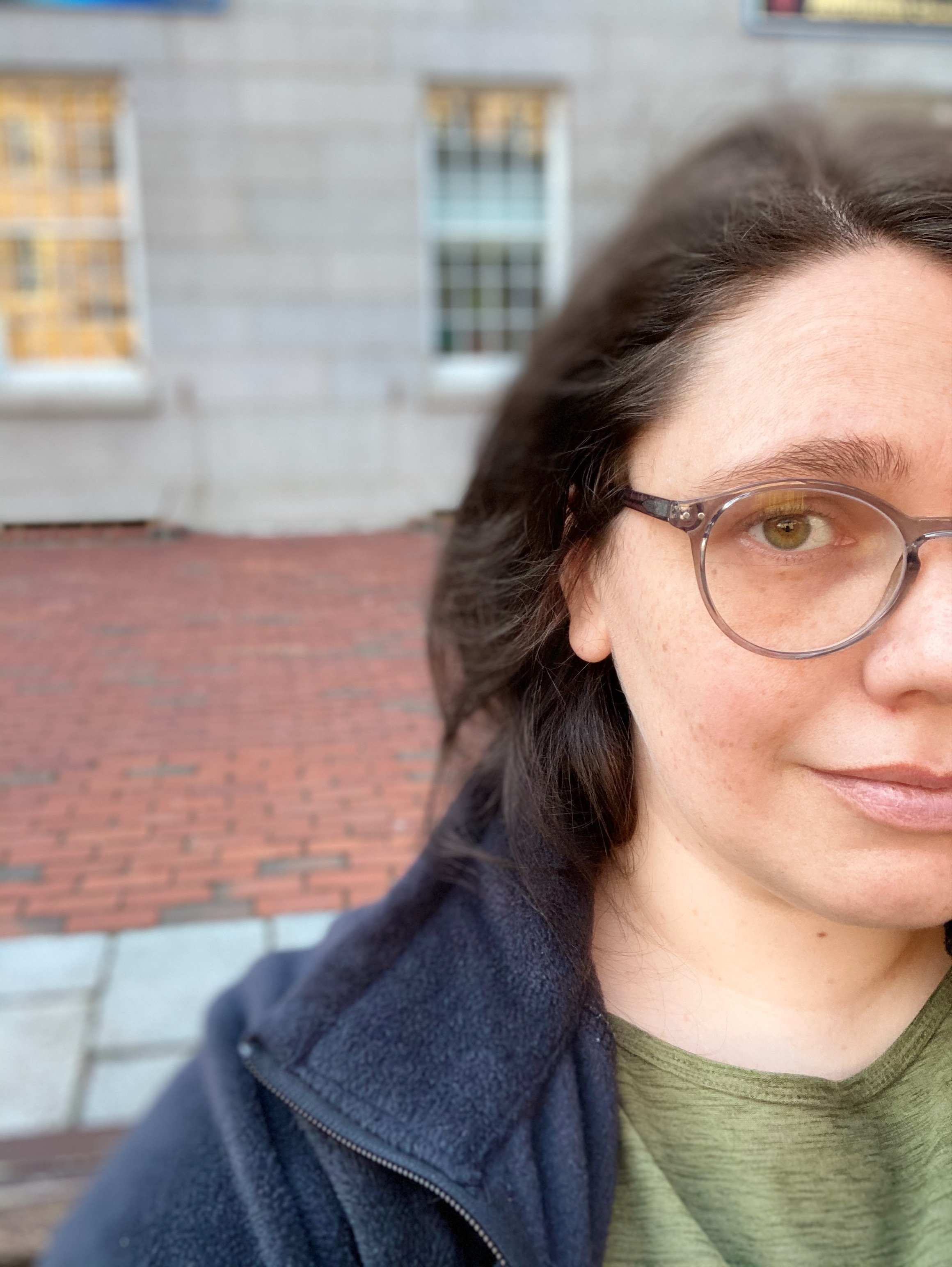 Close-up portrait of a woman wearing glasses and a navy jacket in front of a brick-and-glass building