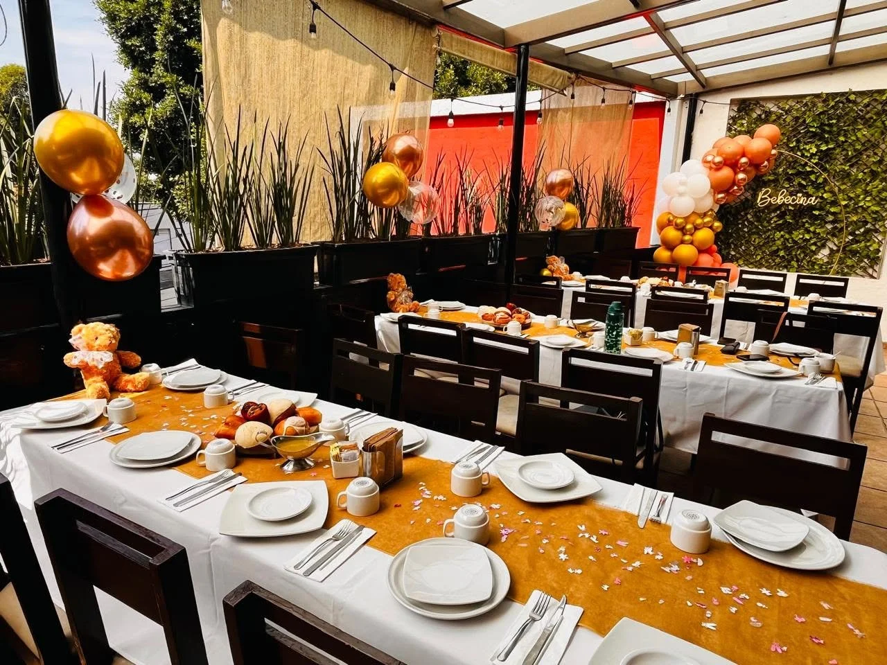 Mesa decorada para celebración con globos de colores metálicos y blancos, un muñeco de felpa, confeti en forma de estrellas y flores, y un fondo con plantas y decoración de globos.