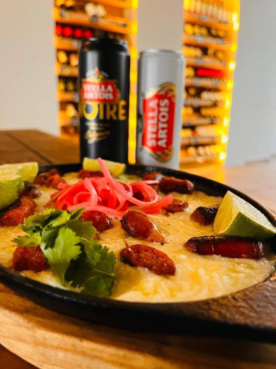 Plato de queso fundido con chorizo y guarniciones de limón, cebolla encurtida y cilantro, en una mesa de madera, con latas de cerveza en el fondo.