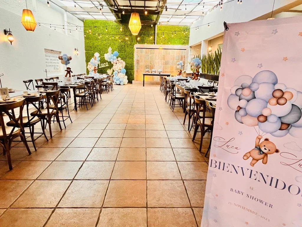 Decoraciones para un baby shower con tema de ositos y globos, mesas arregladas para una fiesta, cartel de bienvenida con el nombre Luca y fecha del evento, globos en tonos pastel y plantas decorativas.