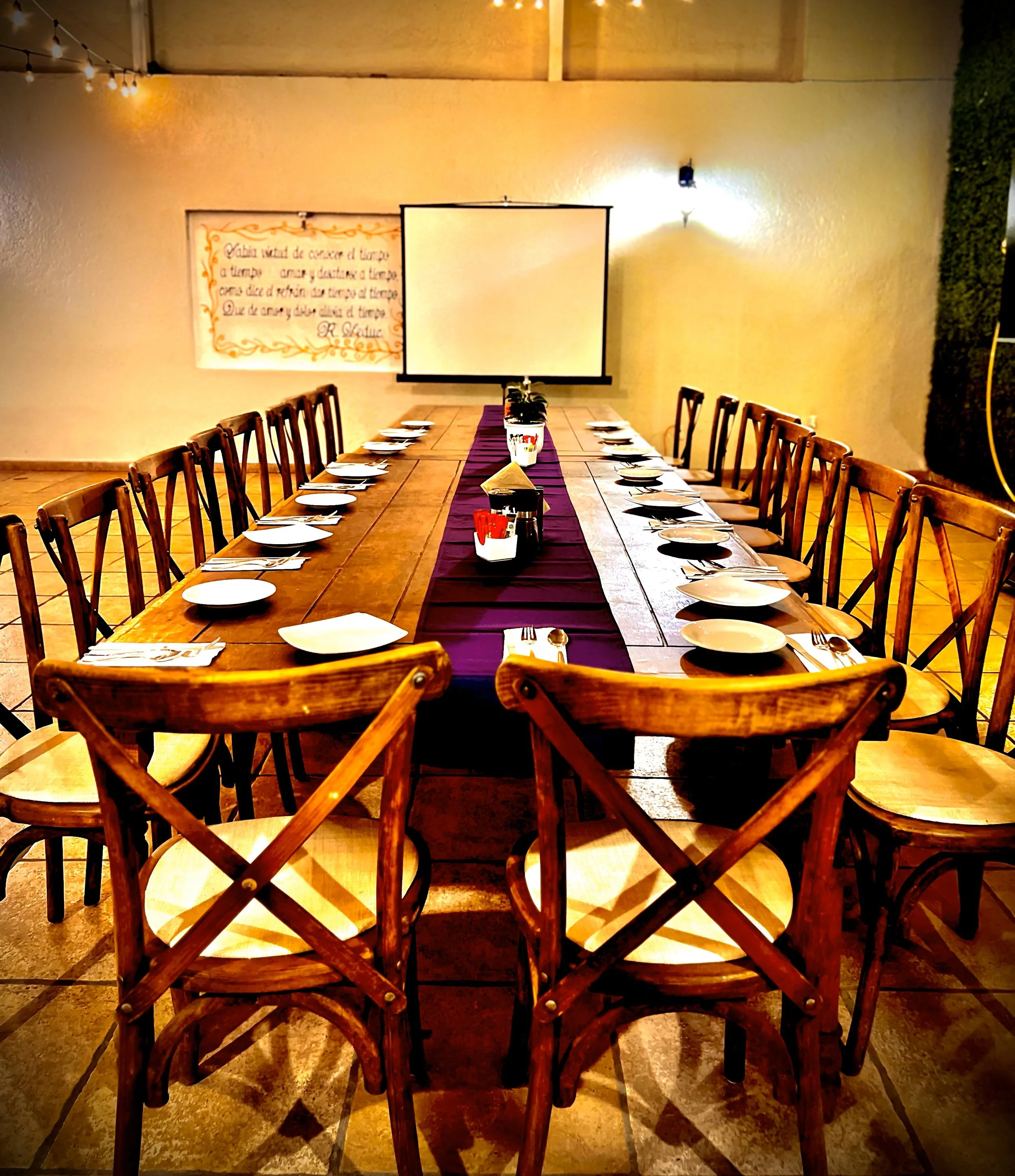 Mesa larga de madera en un comedor con sillas de madera, decorada con un camino púrpura y vasos y platos blancos. En la pared, hay un pizarrón blanco y un cartel decorativo con escritura en español. Luz cálida y una pared con vegetación en el fondo.