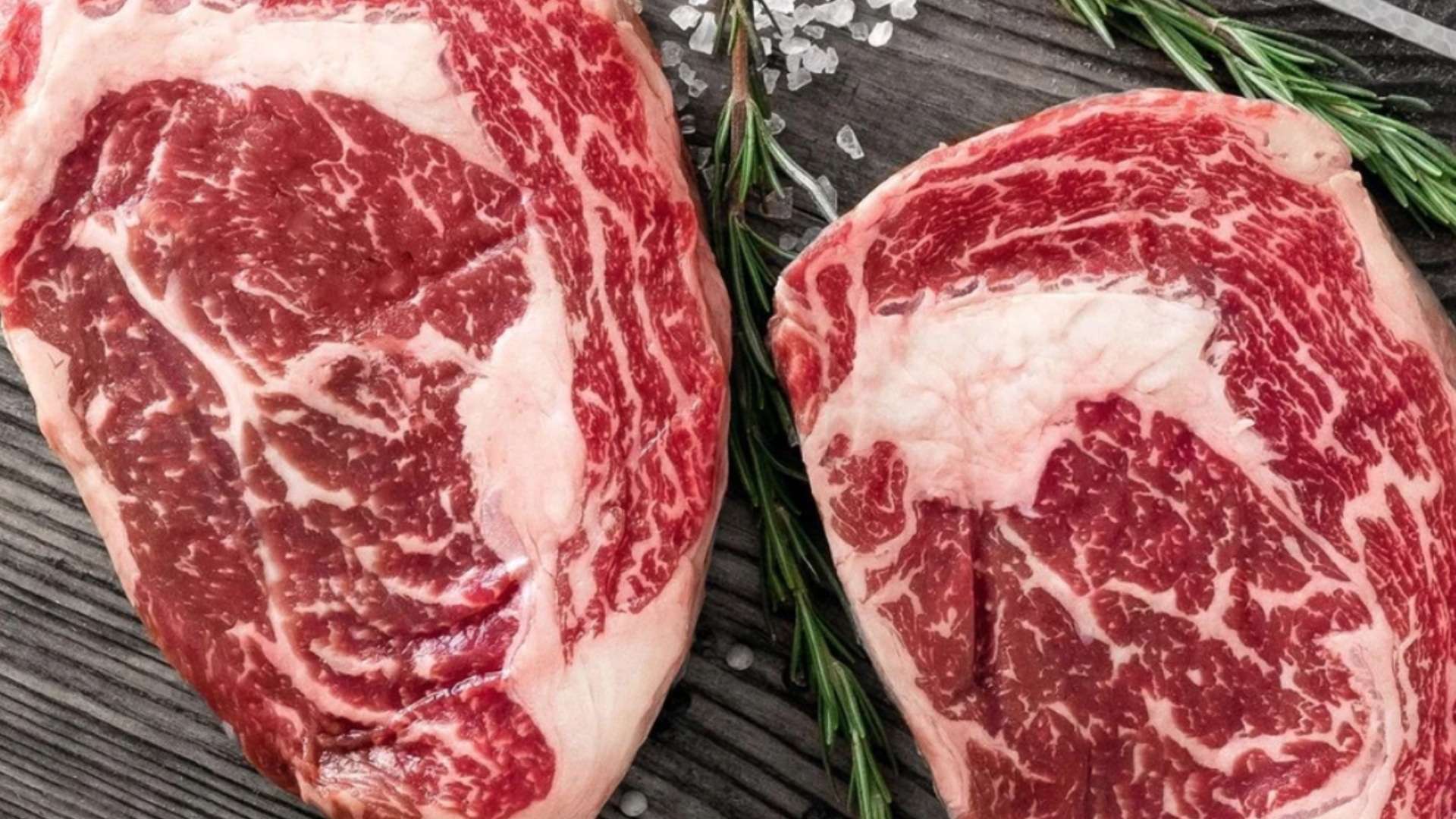 Dos cortes de carne de res con vetas de grasa y romero en una tabla de madera
