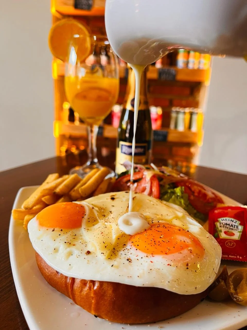 Desayuno con huevos fritos sobre un pan, acompañados de tocino, ensalada, salsa, y cereal, con una bebida en un vaso al fondo.