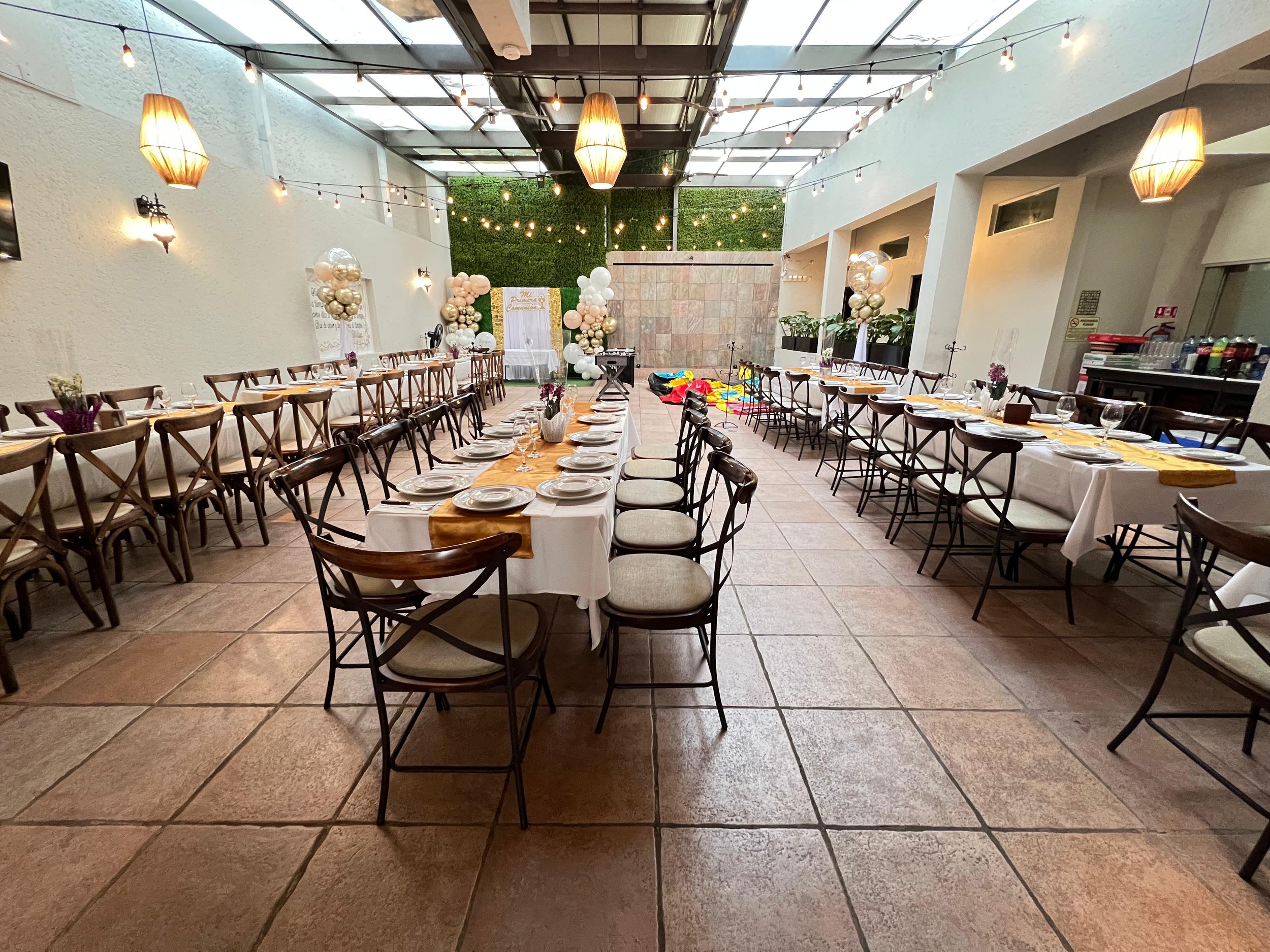 Sala decorada para una celebración o evento, con mesas arregladas con manteles blancos y caminos dorados, decoraciones con globos de colores pastel y metálicos, y globos en forma de columnas, en un espacio cerrado con techo de vidrio y paredes blancas.