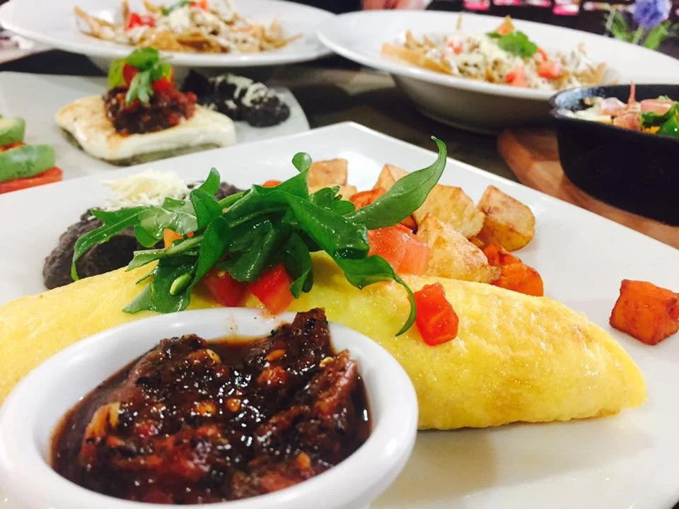 Plato de desayuno con omelette, salsa, verduras y papas, acompañado de ensaladas y otros platillos en fondo.